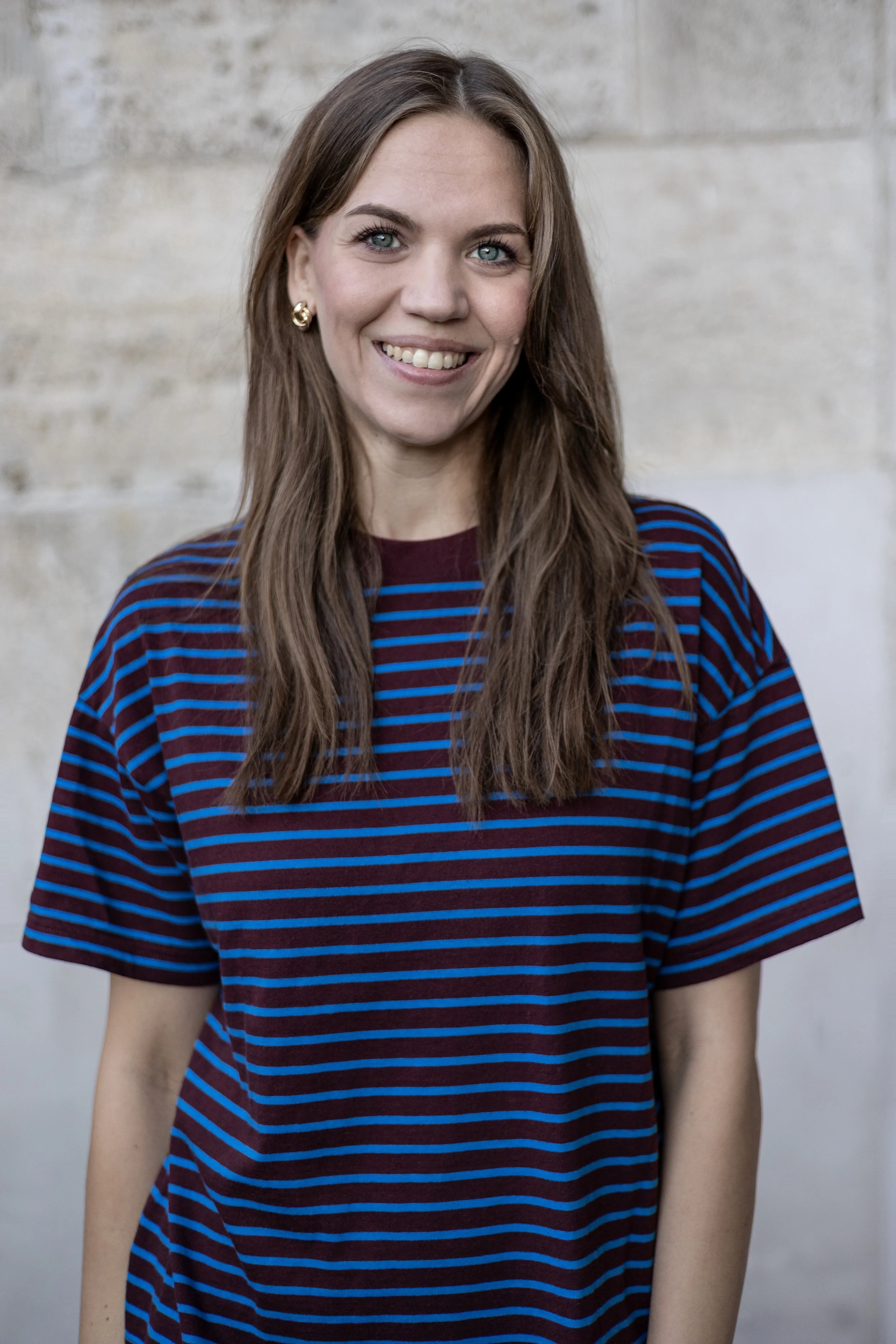 Ein junges Mädchen mit langen braunen Haaren, blauen Augen und einem freundlichen Lächeln, trägt ein rotes T-Shirt mit blauen Streifen, vor einer grauen Steinmauer.