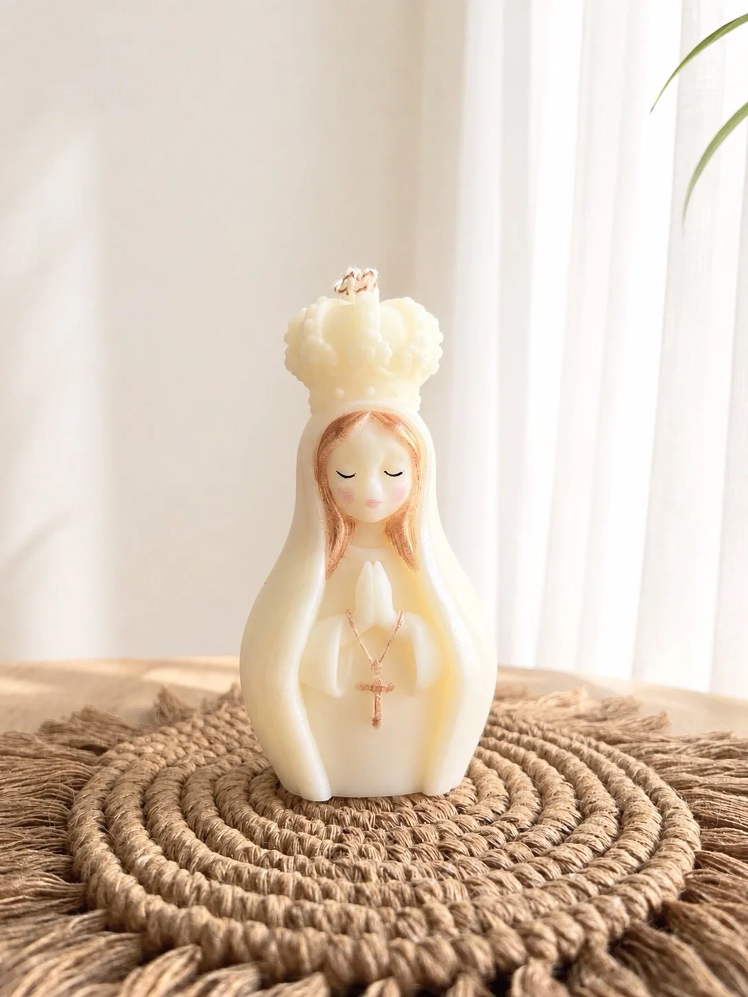 Vela artesanal de una virgen con manos en posición de oración, con un rosario colgado y una corona en la cabeza, en una base de macramé sobre una superficie de madera.