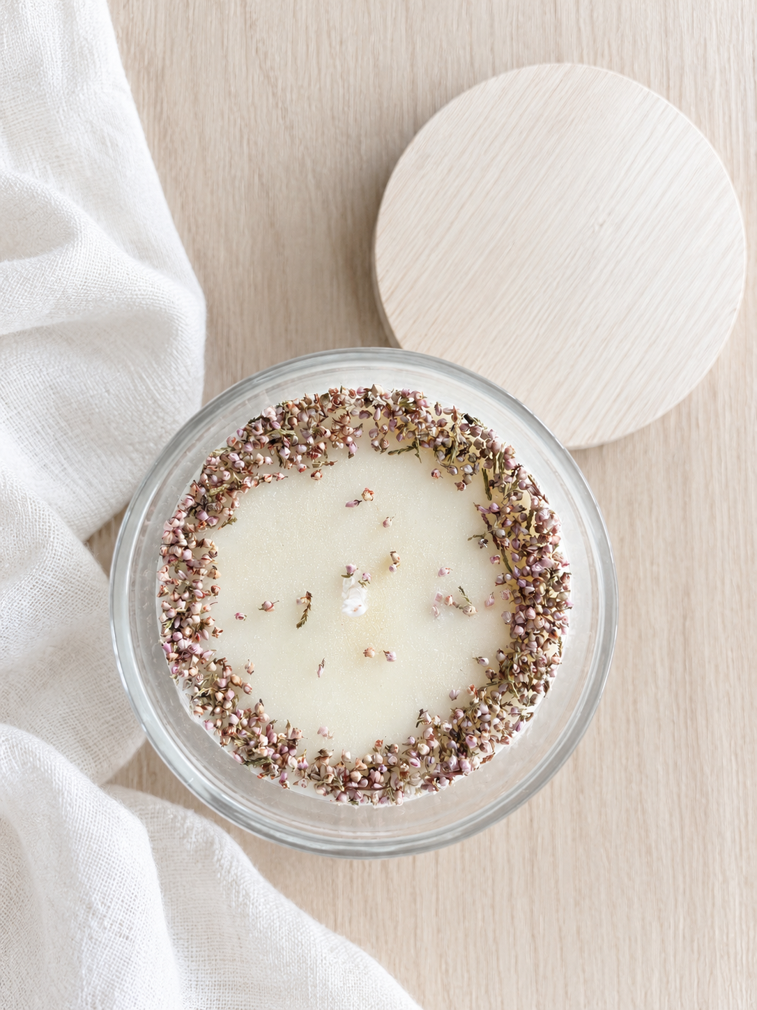 Vela de cera blanca en frasco de vidrio, decorada con flores secas en el borde, sobre superficie de madera clara con una tapa de color madera claro y pedazo de tela blanca a la izquierda.