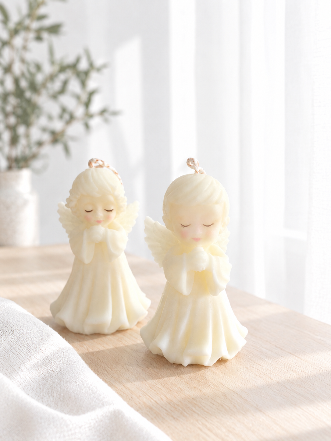Dos velas en forma de ángeles con manos en posición de oración, decoradas con gracia, en un entorno iluminado con luz natural.
