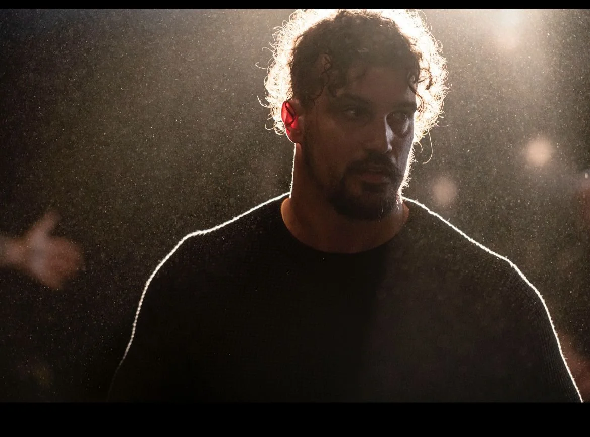 A man with curly hair and facial hair, wearing a dark shirt, is backlit with a bright light creating a halo effect and highlighting particles or dust in the air.