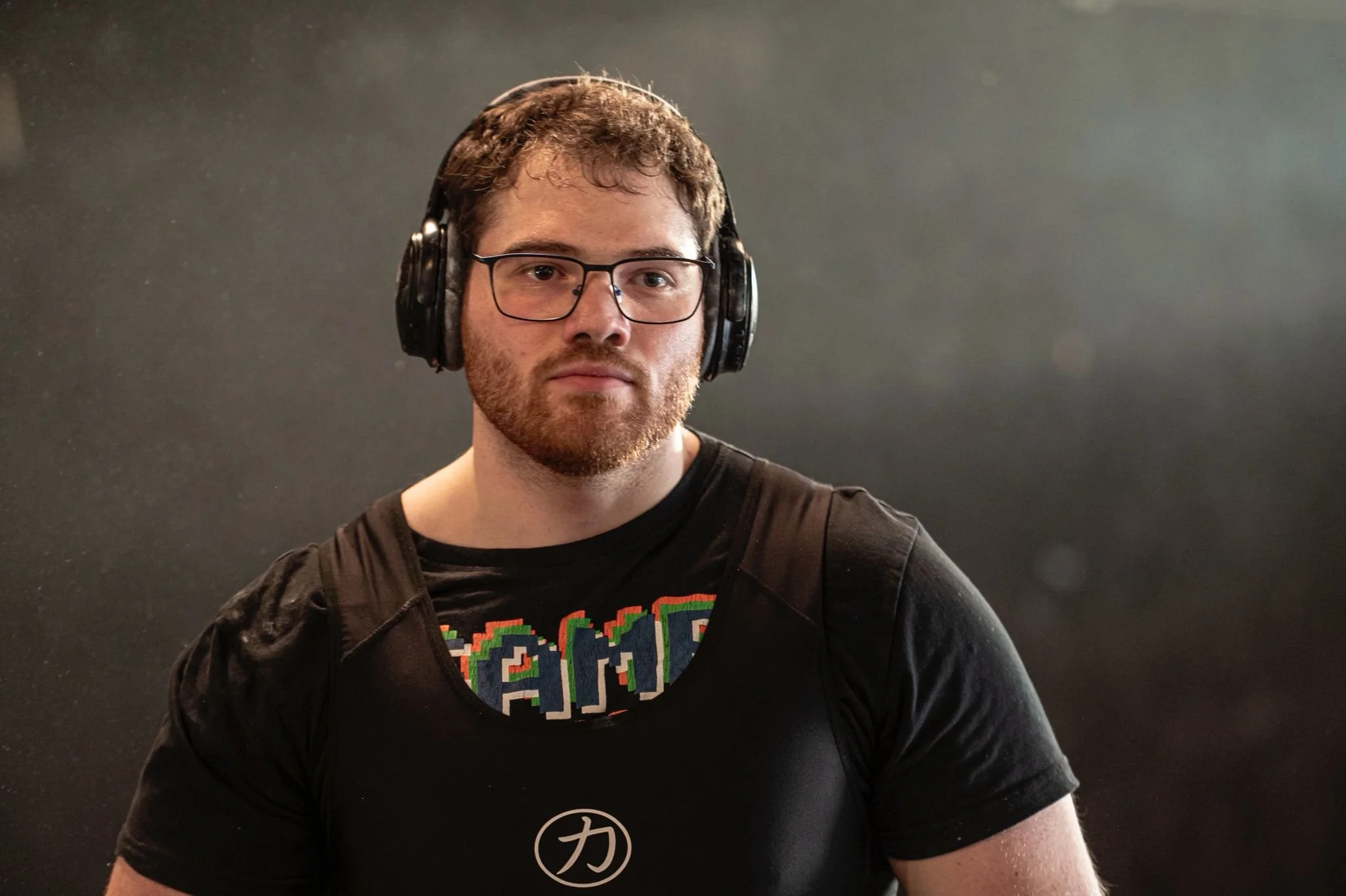 A man with glasses and a beard wearing headphones, a black t-shirt with colorful text, and a backpack against a dark background.