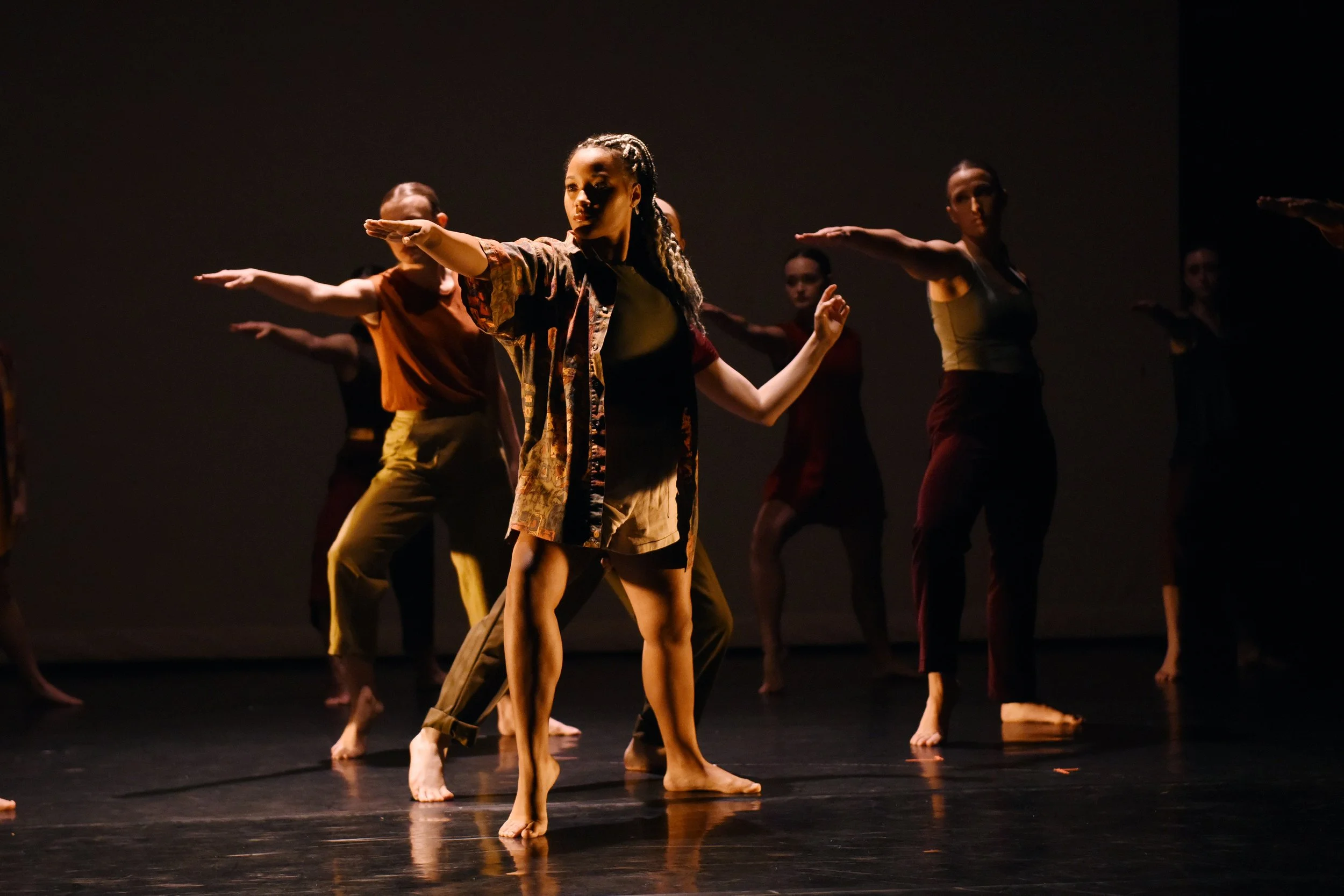 Group of dancers performing on stage, with one woman in the foreground extending her arm and looking to the side, barefoot, wearing a patterned jacket and shorts.
