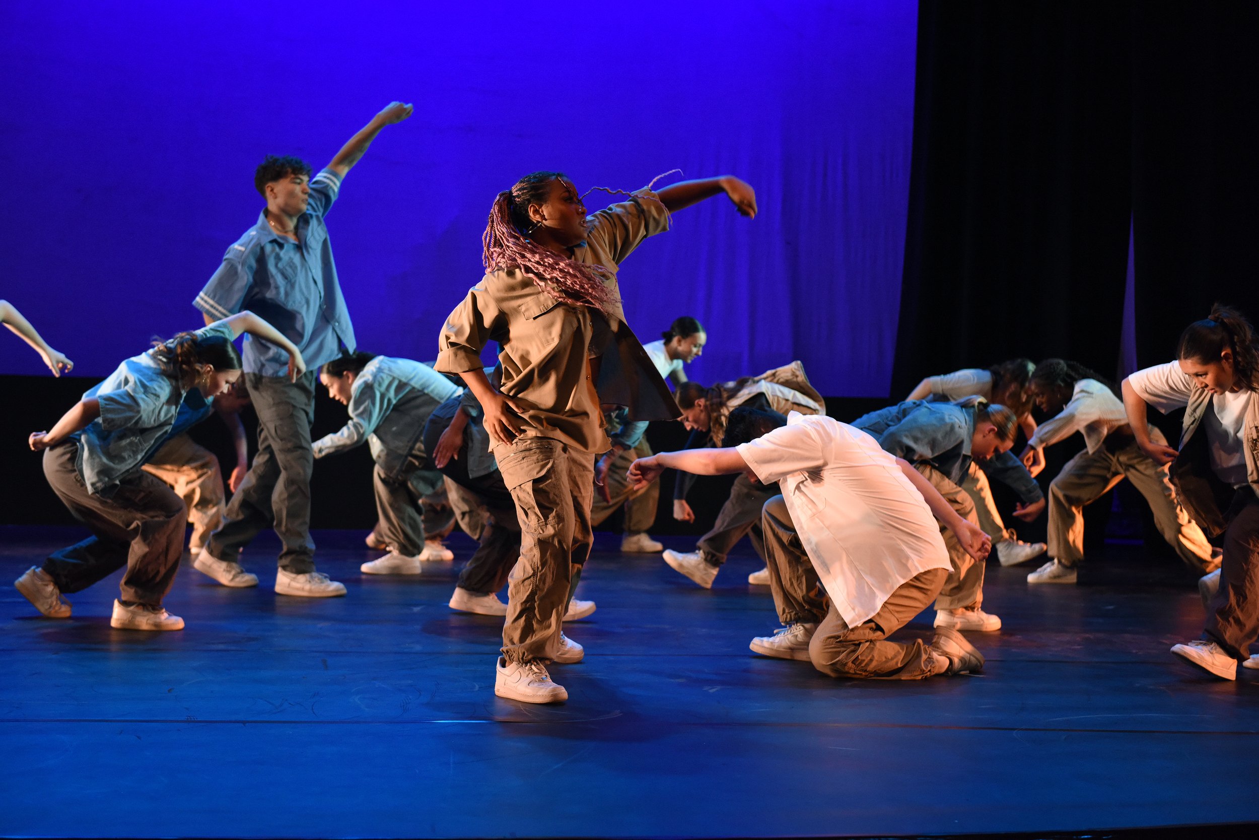 A group of dancers performing on stage with a blue background, wearing casual outfits including khaki pants and topped T-shirts, captured mid-movement.
