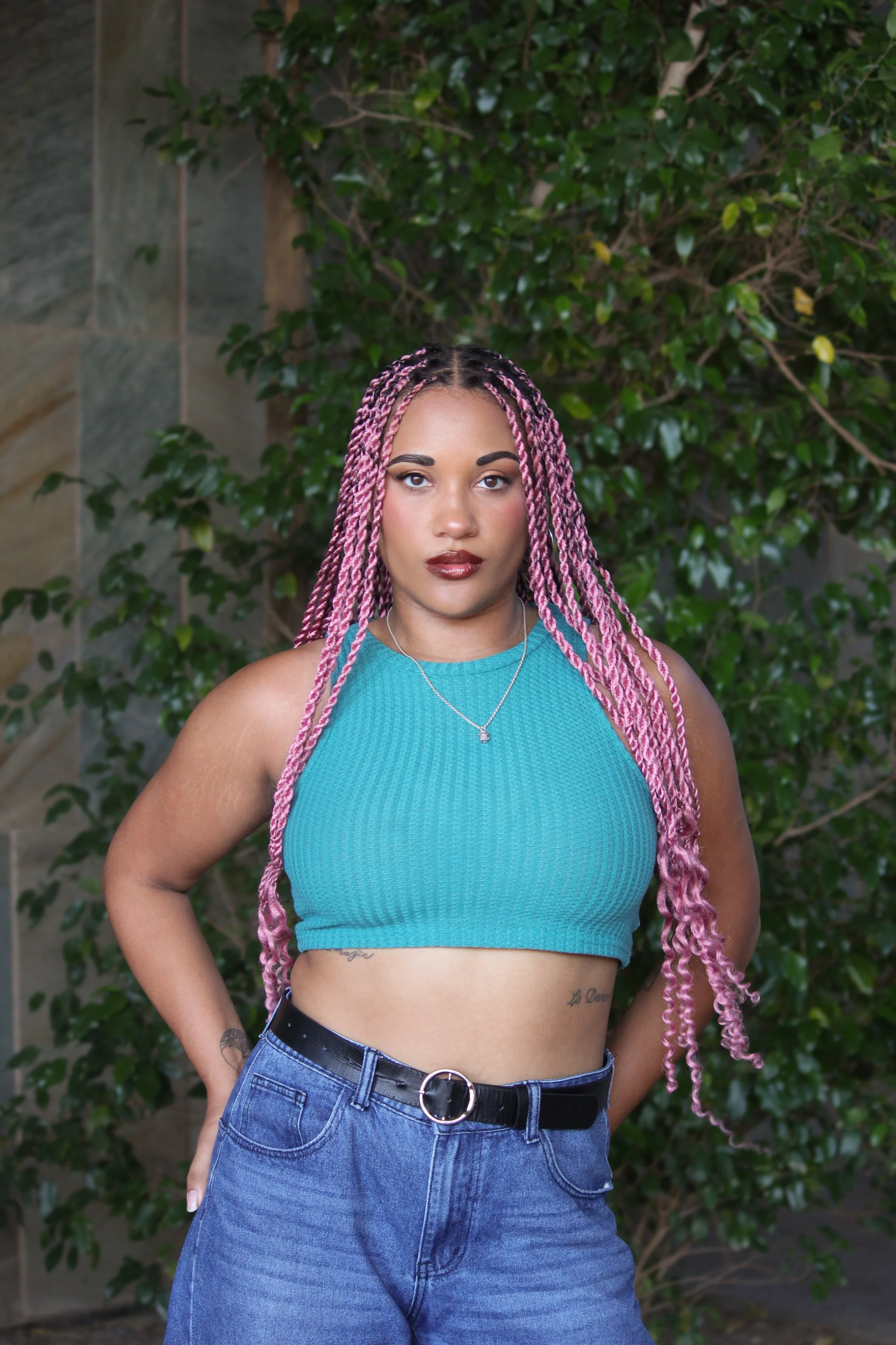 A woman standing outdoors in front of green foliage, wearing a turquoise sports bra, blue jeans, and a black belt, with long pink twists.