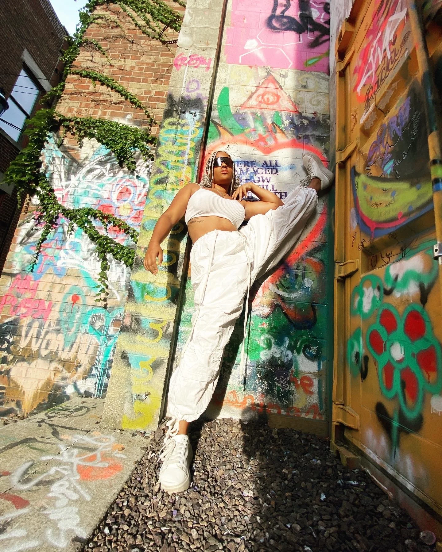 A woman with braided hair, wearing sunglasses, a white crop top, white baggy pants, and white sneakers, is leaning in a split against a graffiti coverd wall in an alley.