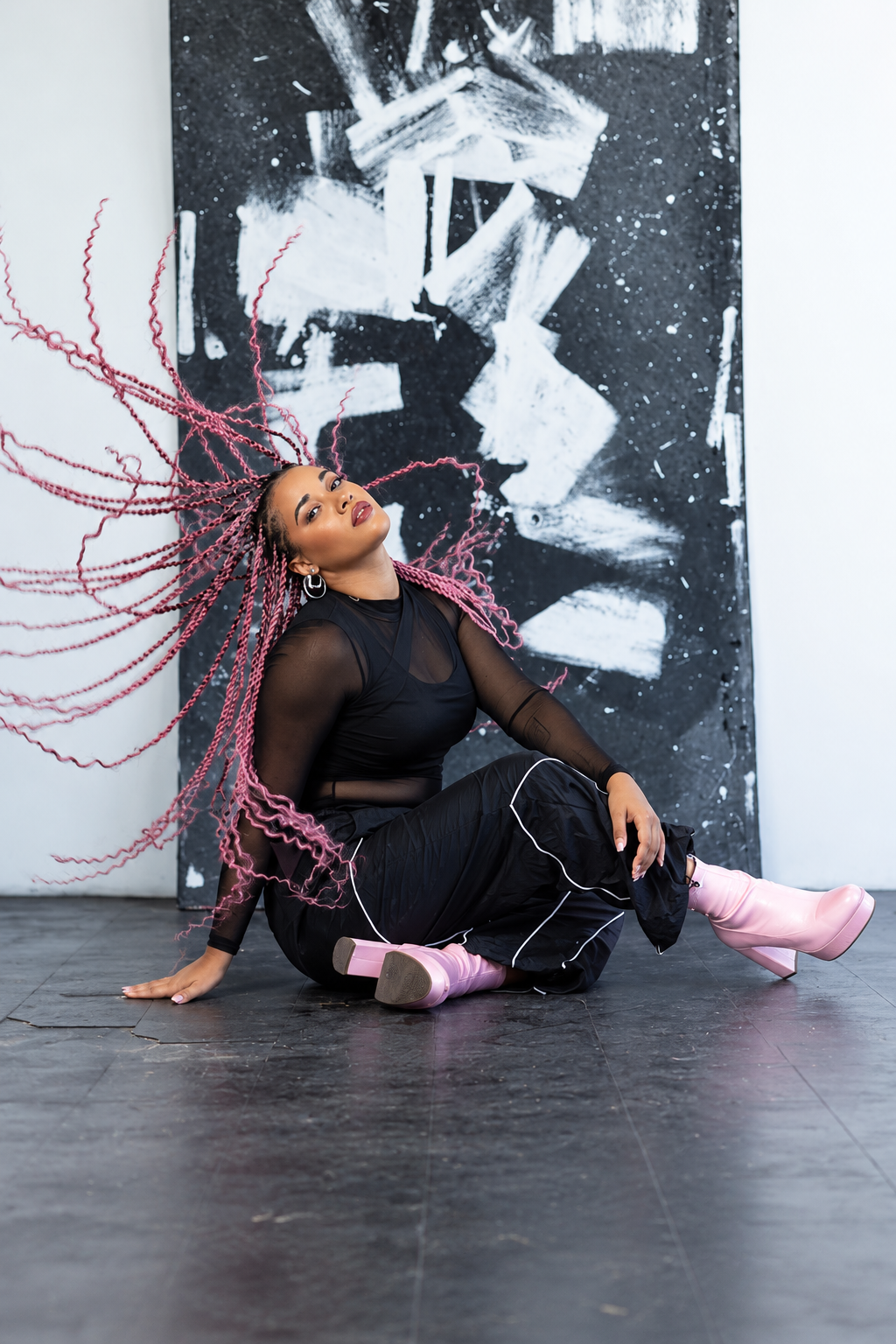 A woman with long pink twists and hoop earrings poses on the floor in front of abstract black and white art, wearing a black sheer top, black pants with white piping, and pink platform boots.