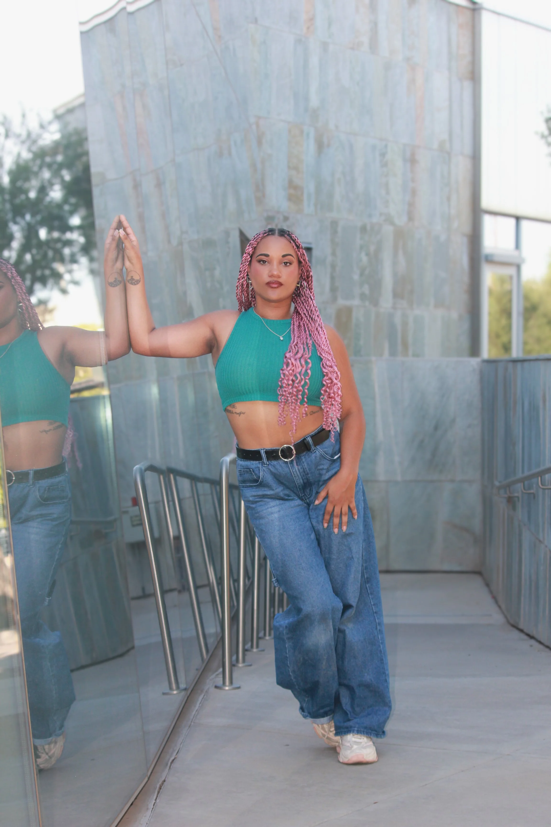 A woman with long pink twists in a teal crop top and blue jeans is standing outside near a glass wall, touching her reflection with her right hand, and looking at the camera.
