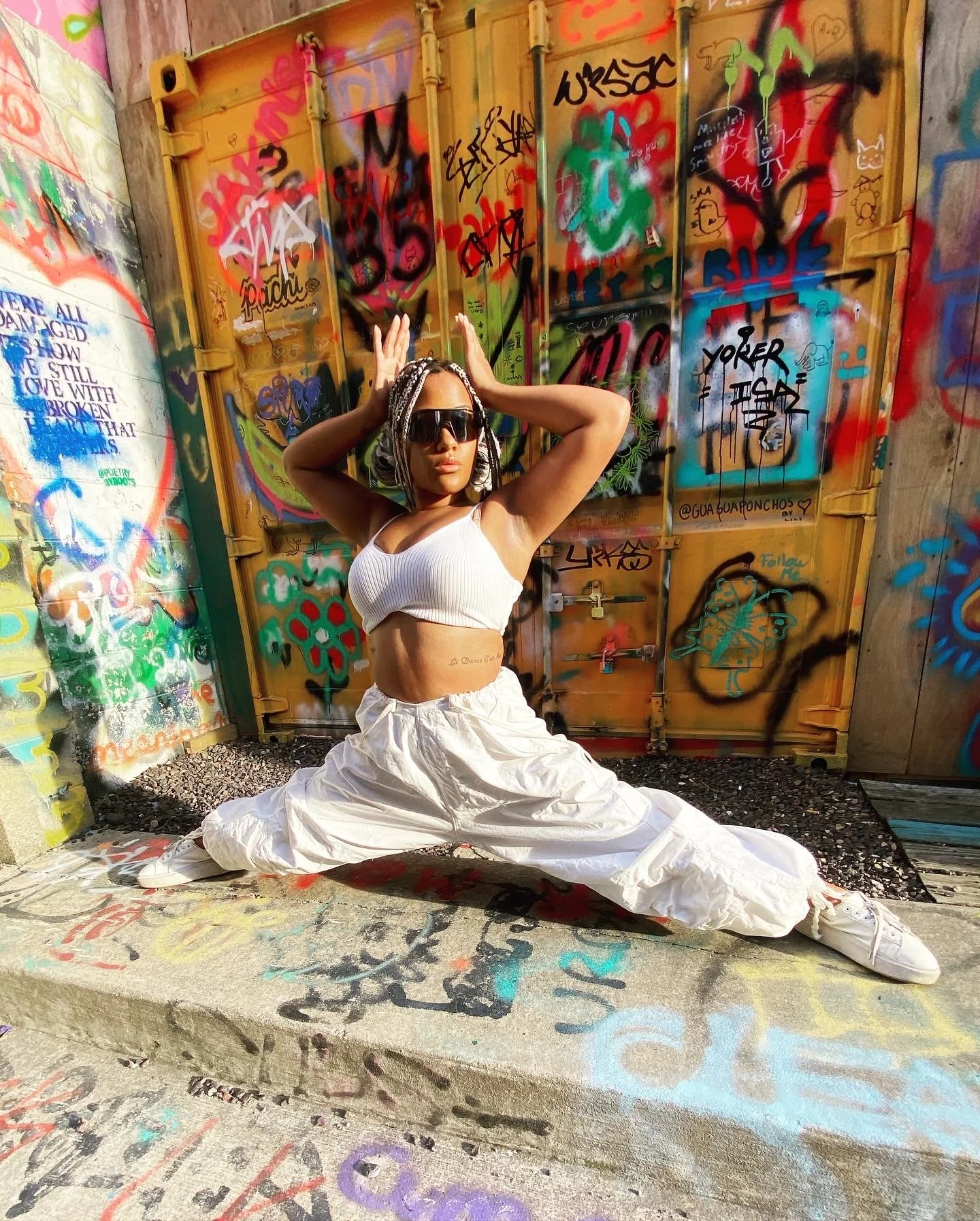 A woman with braided hair, wearing sunglasses, a white crop top, and white baggy pants, poses in a deep center split. She has her hands raised above her head in front of a background of colorful graffiti-covered metal containers.