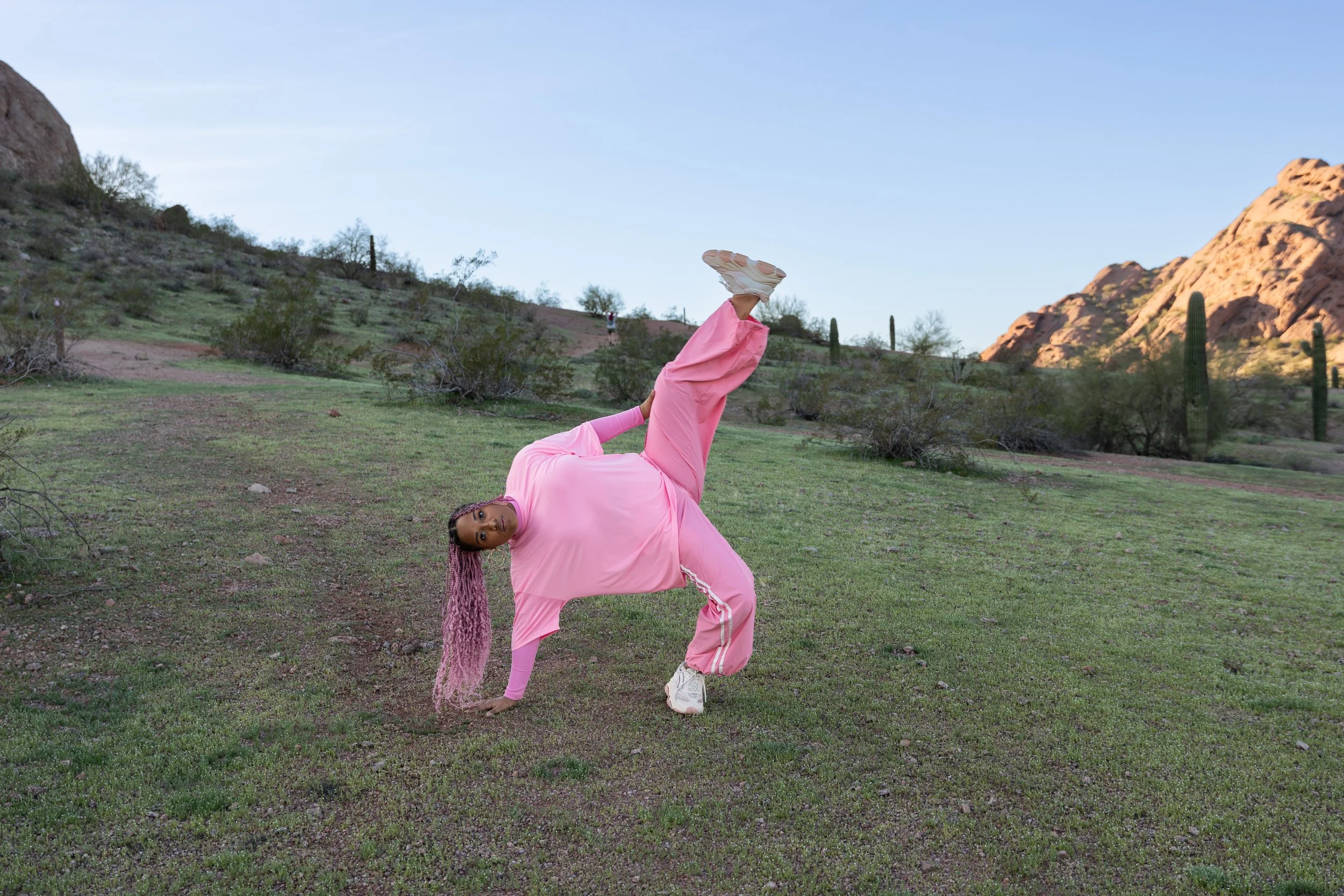 A woman in a pink t-shirt and pants performing a backbend on one hand with her leg outstretched in the air outdoors in a desert landscape with mountains, cacti, and sparse vegetation.