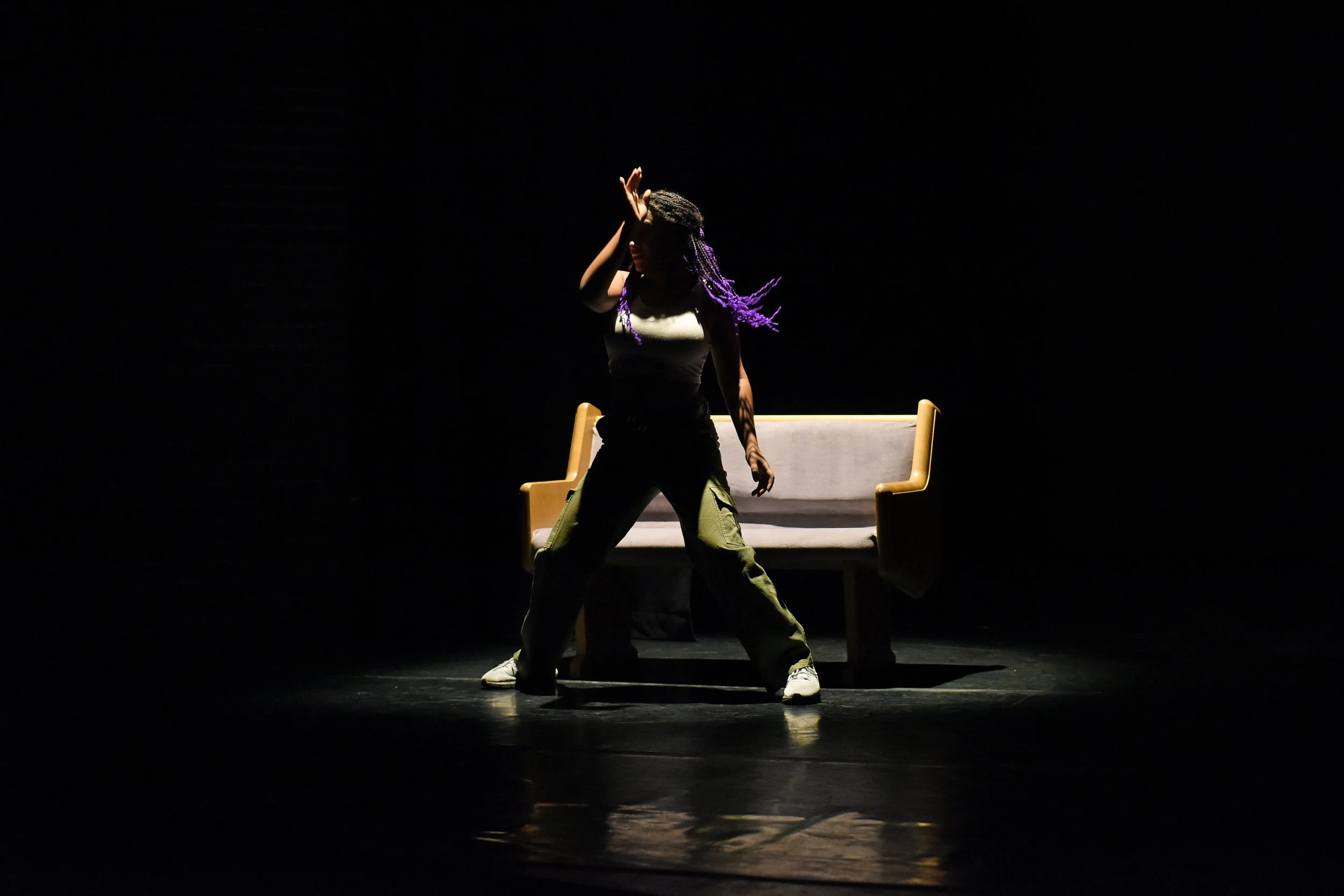 A woman with purple braided hair standing in a spotlight on stage, with a bench behind them. The background is dark.