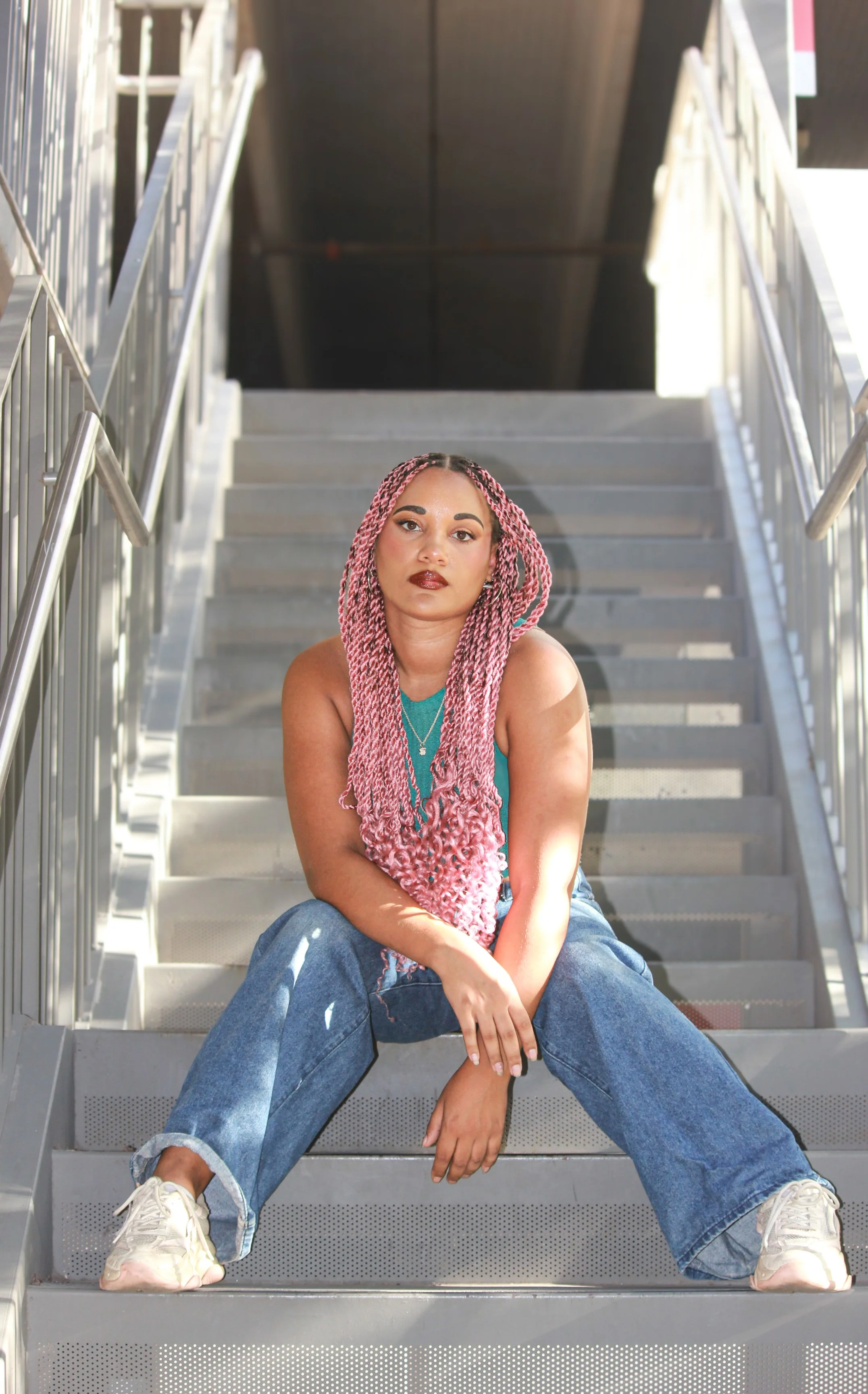 Young woman with long pink twists sitting on outdoor metal stairs, wearing a teal top, blue jeans, and white sneakers, posing confidently.