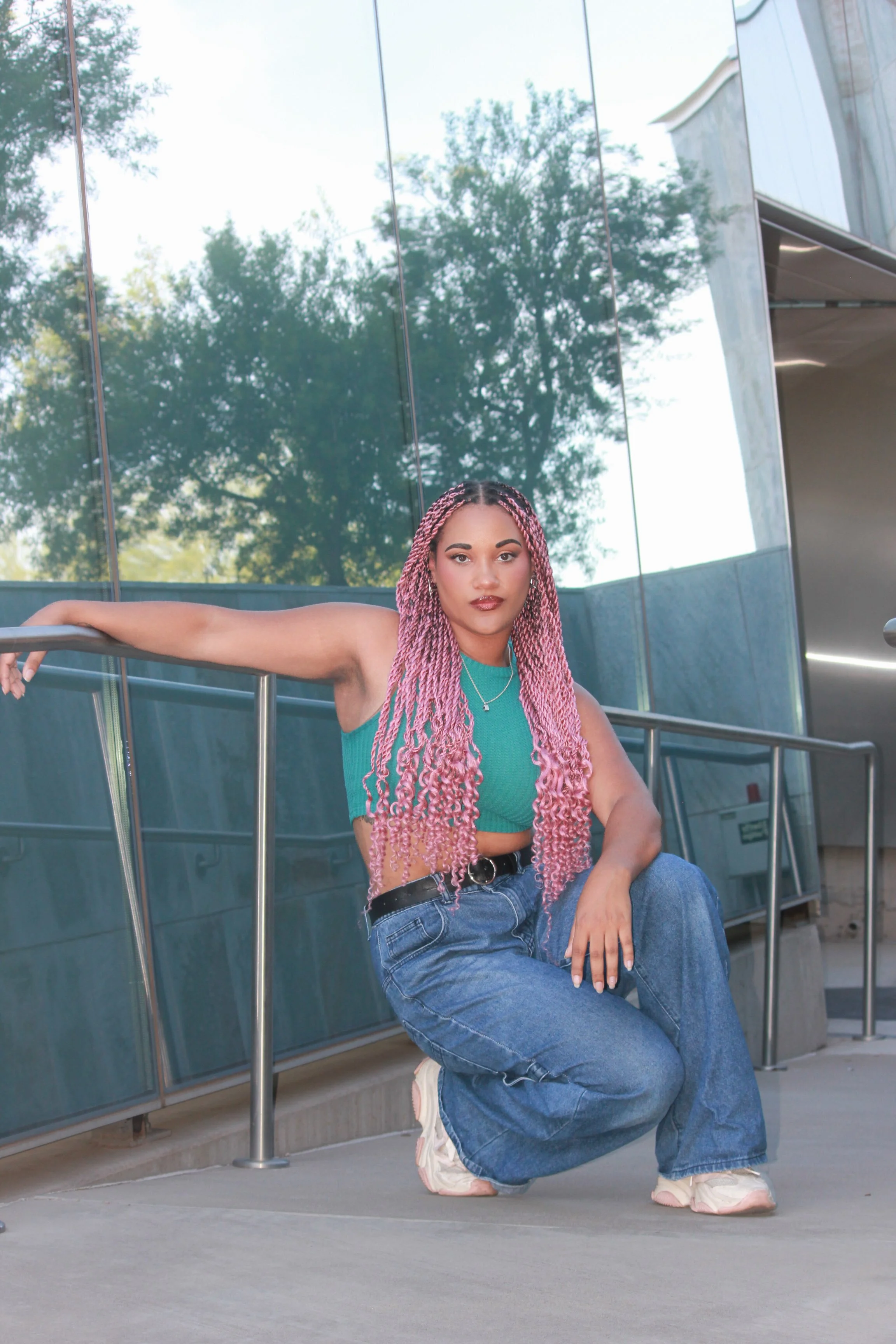 Young woman with long pink twists, wearing a teal tank top and baggy blue jeans, crouching outdoors near a metal railing with a glass building and trees in the background.