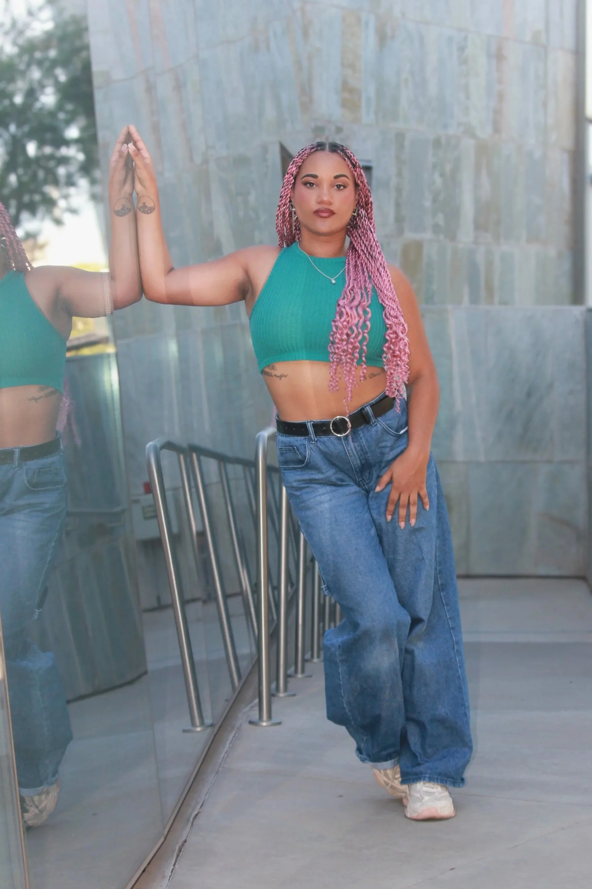 A young woman with long pink twists, wearing a teal crop top and baggy jeans, is standing indoors next to a glass window, with her hand pressed against the glass in a mirror reflection.