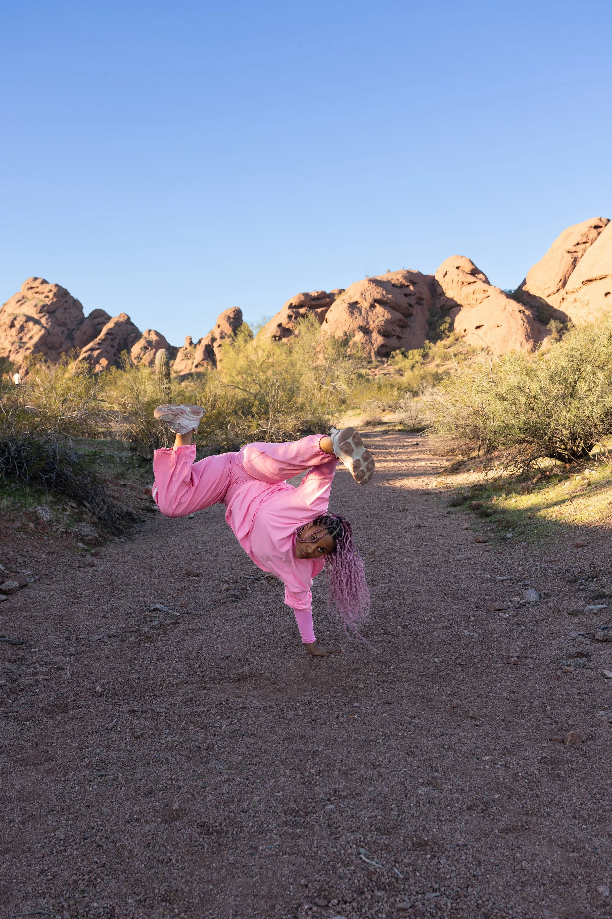 A woman with pink hair wearing a pink t-shirt and pants doing a k-stand on a dirt path in a desert landscape with rocky hills and sparse desert vegetation under a clear blue sky.