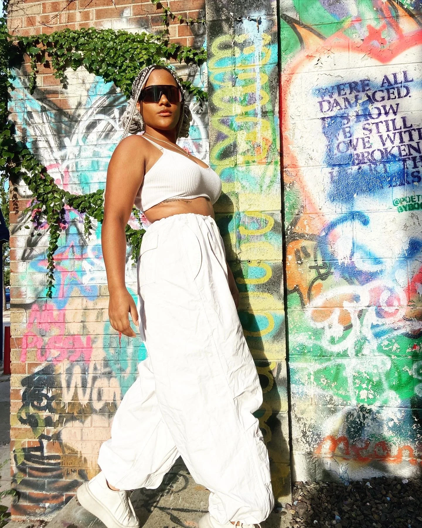 Woman in white crop top, baggy white pants, white sneakers, and oversized sunglasses standing against a colorful graffiti wall with poems and art, partially covered by green ivy.