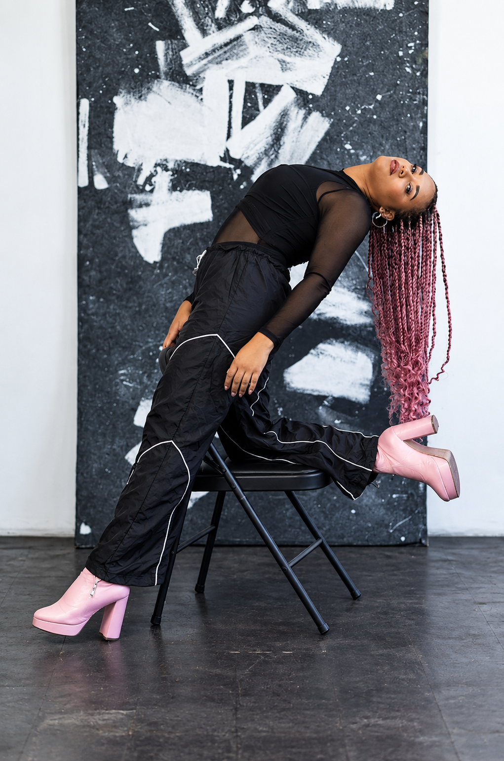 A woman with long pink twists leaning backward, one leg resting on a chair and the other extended, wearing black sheer top, black pants with white piping, and pink high-heeled boots, in front of an abstract black and white painting on a white wall.