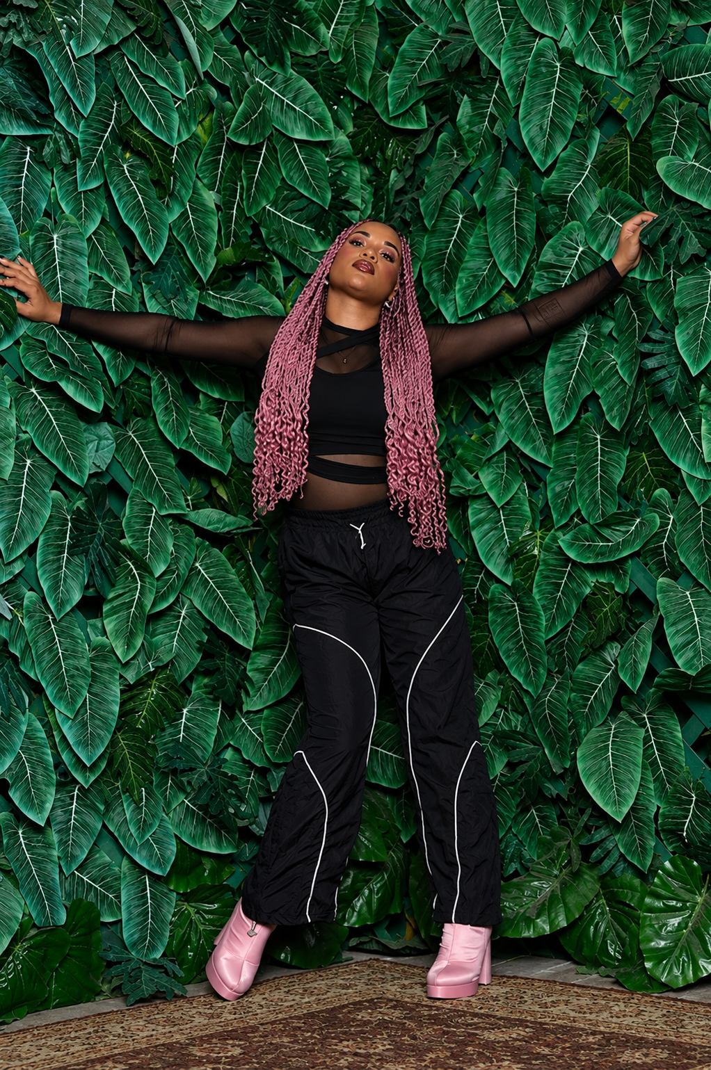 A woman with long pink twists poses with her arms outstretched in front of a green leafy wall. She wears a black sheer long sleeve top, black pants with white piping, and pink platform boots.
