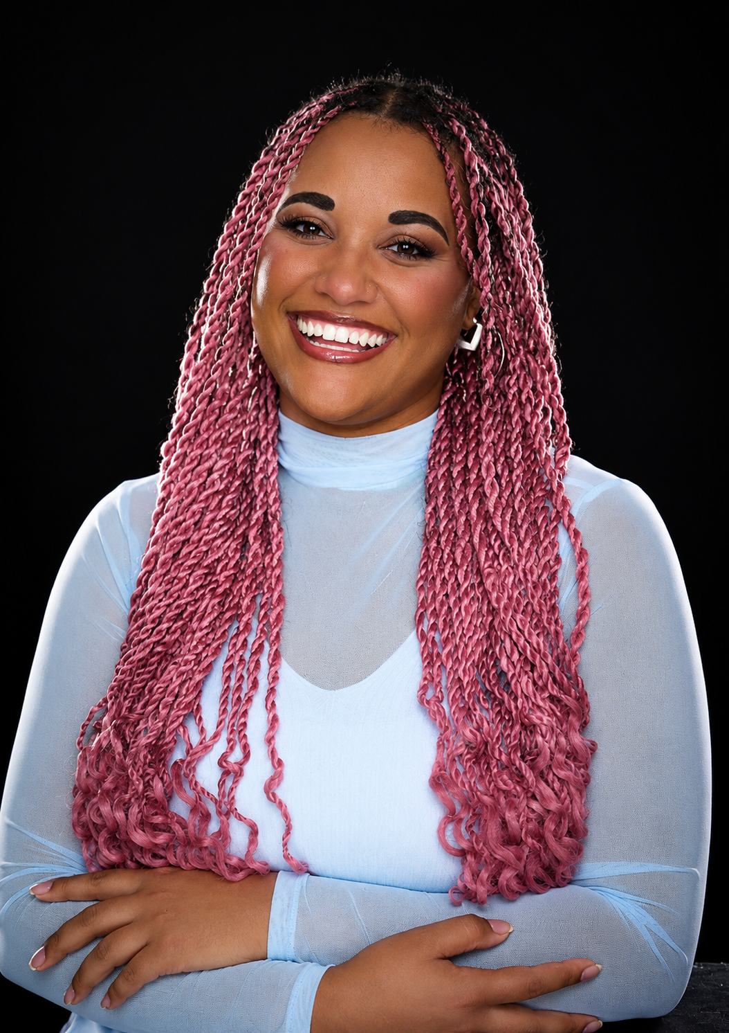 A smiling woman with long pink twists, wearing a light blue long-sleeve top and jewelry, on a black background.