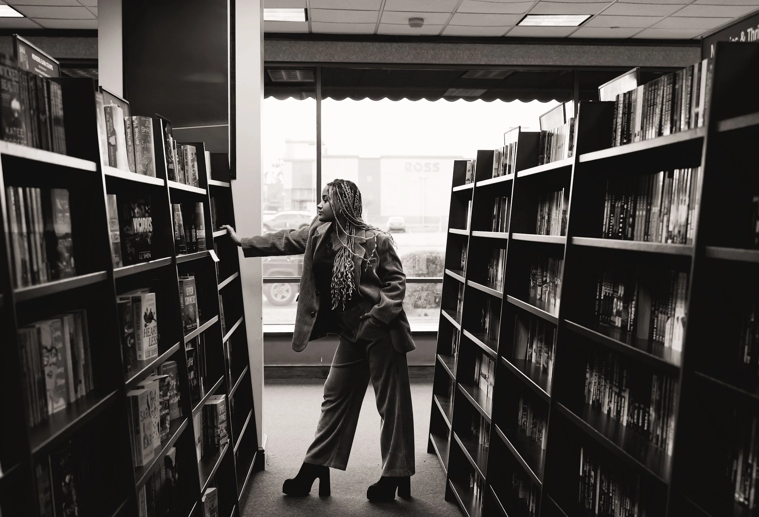 A woman with braids and a coat browsing books in a bookstore aisle, with sunlight coming through the window behind her.