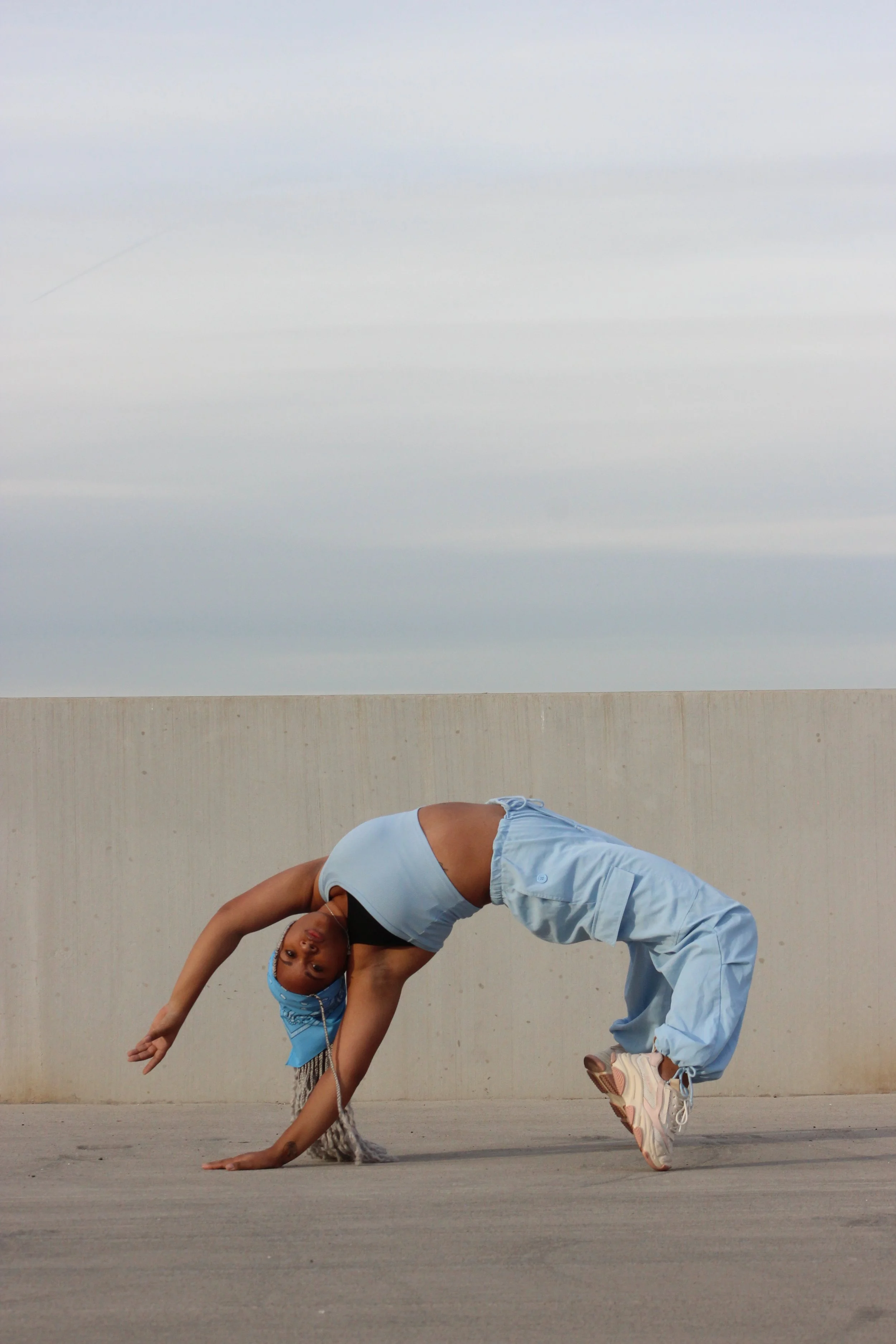 A young woman dressed in light blue athletic clothing and a matching headband performing a dance move in front of a plain wall, with her body arched backward and one hand touching the ground.