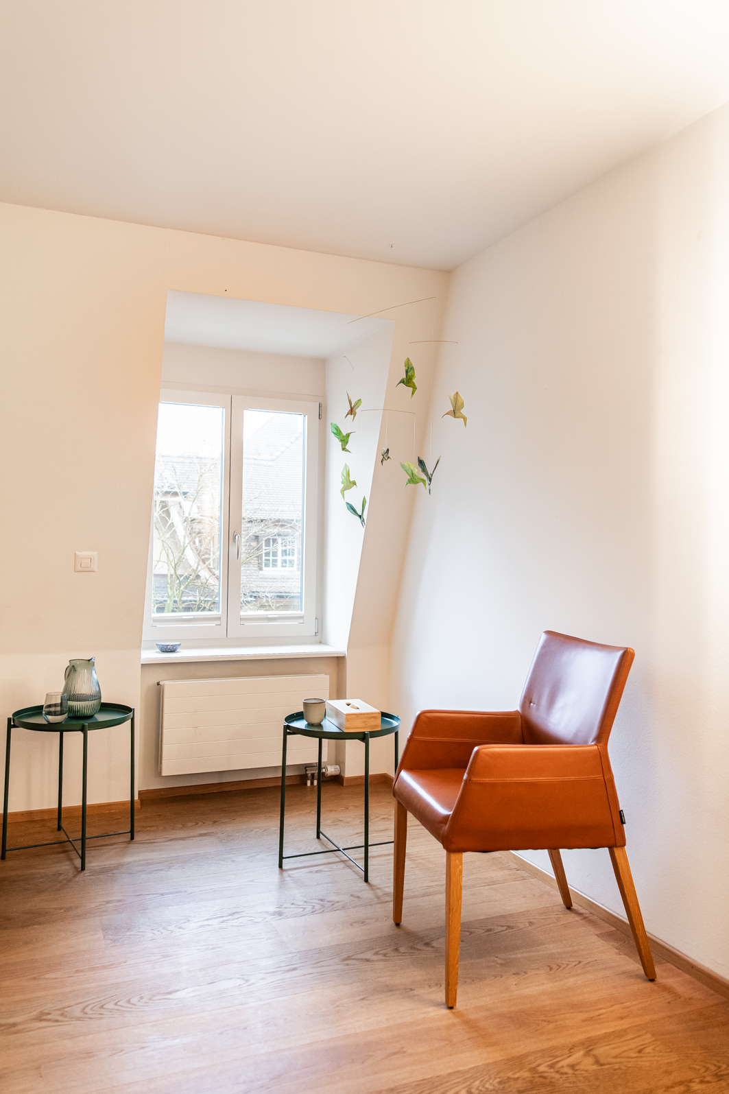 A cozy corner of a room with light-colored walls, a window with a white frame, and a brown leather armchair. Two small black tables hold decorative items like a jug, a glass, a candle, and a box. Hanging butterfly mobile decor is near the window.
