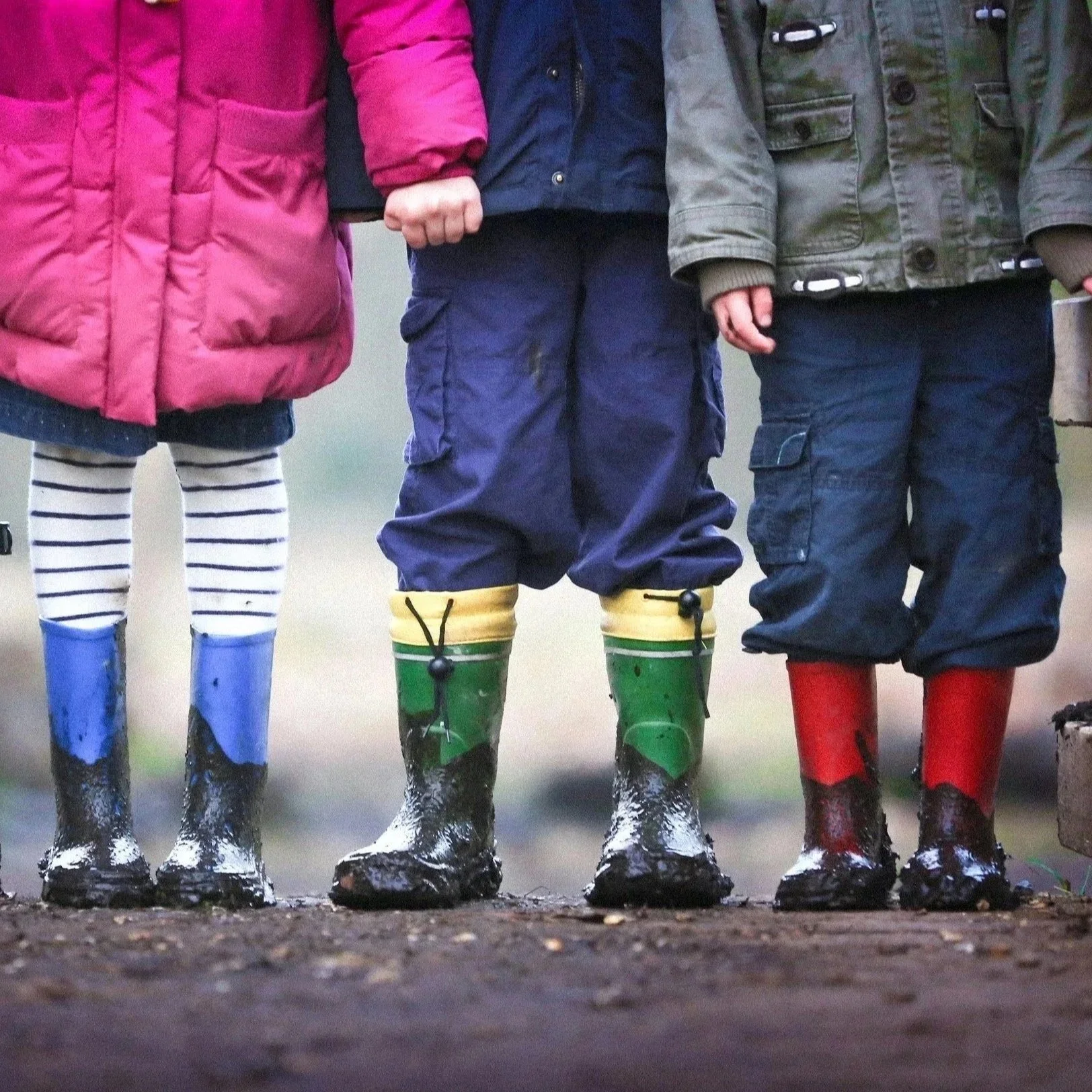 Close-up of four children's feet standing in a row on the ground, wearing colorful rain boots and waterproof clothing, in a muddy outdoor environment.