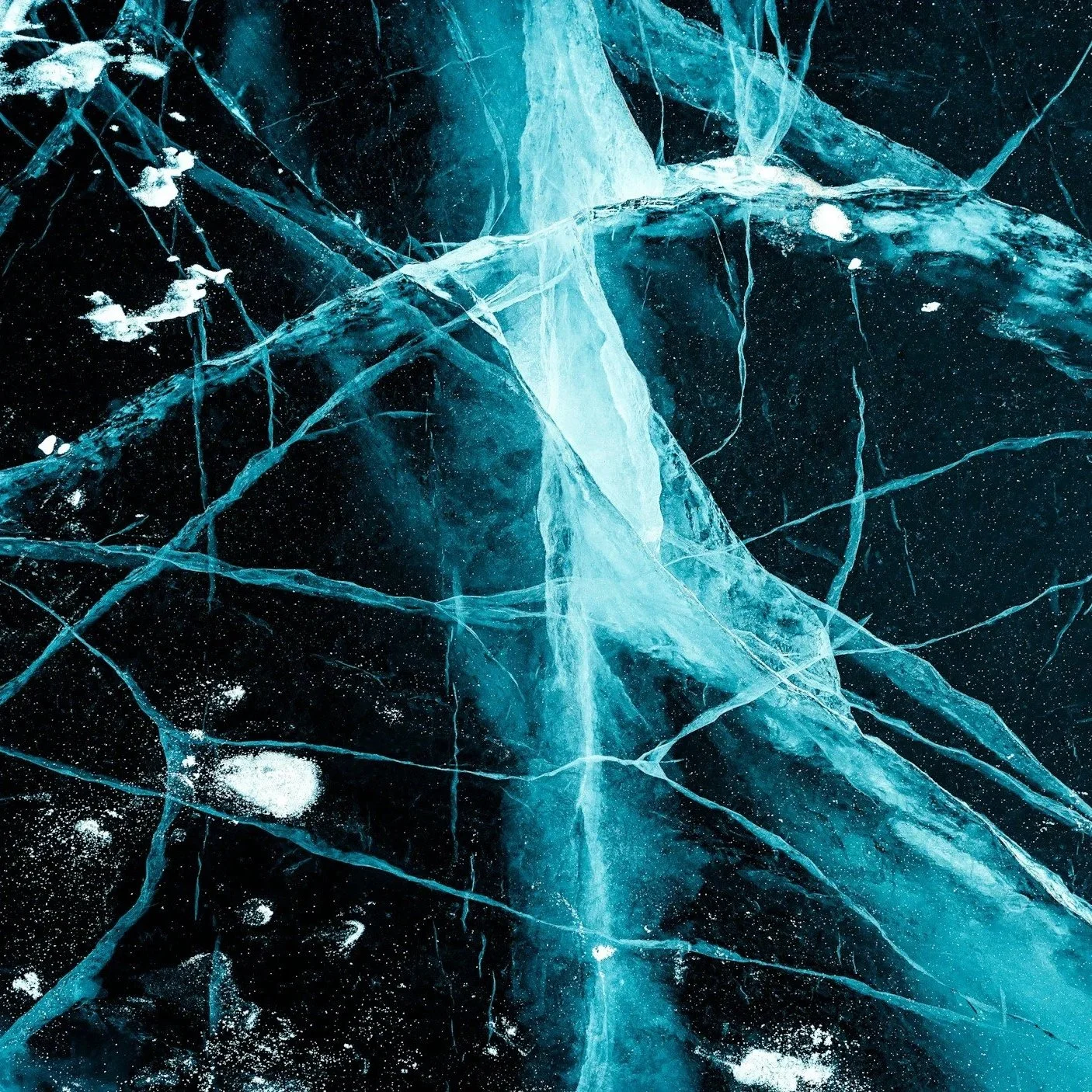 Close-up of a cracked frozen ice surface with blue and black hues, showing intricate fractures and snow dust.
