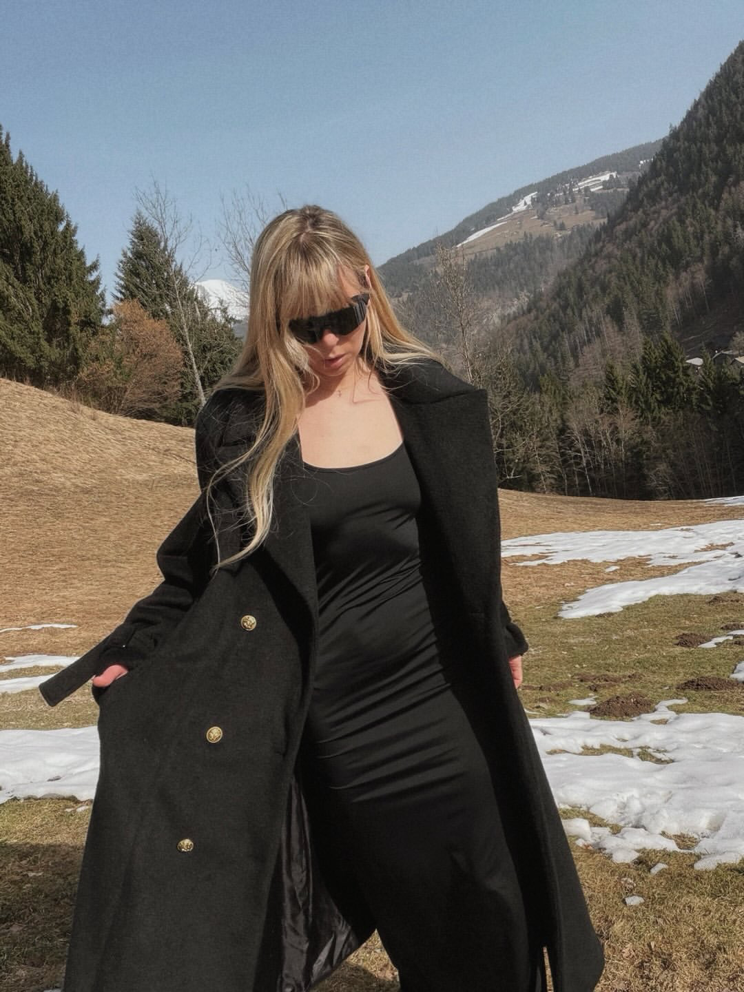 A woman with long blonde hair wearing sunglasses, a black dress, and a black coat standing outdoors in a mountainous area with patches of snow on the ground, trees, and mountains in the background.