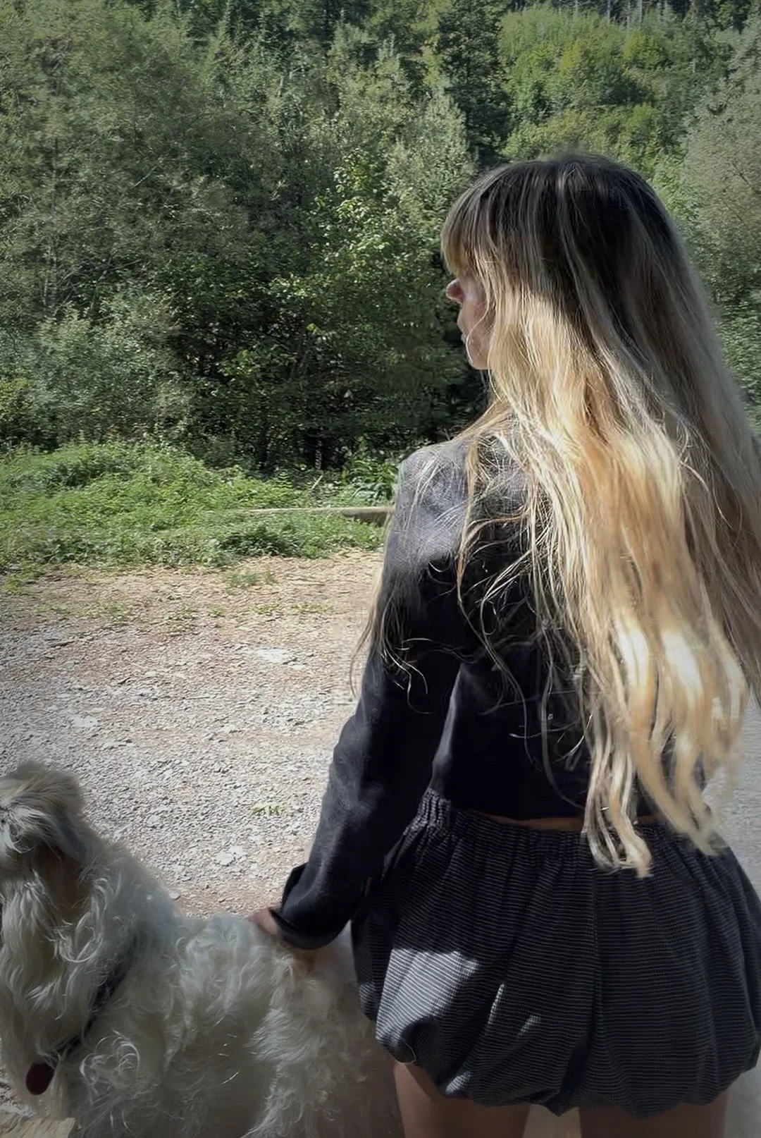 A woman with long, wavy blonde hair standing outdoors with a big white greyhound, a dog, with a dense forest in the background.