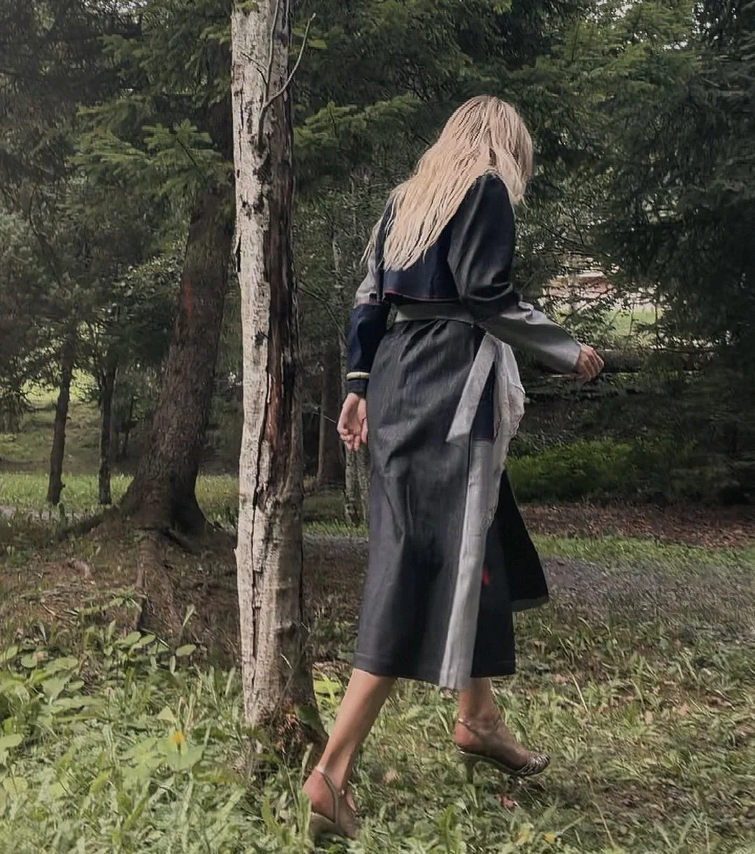 A woman with blonde hair wearing a stylish dark denim and gray patchwork coat, beige handmade high heels, and carrying a handbag, walking in a wooded area.