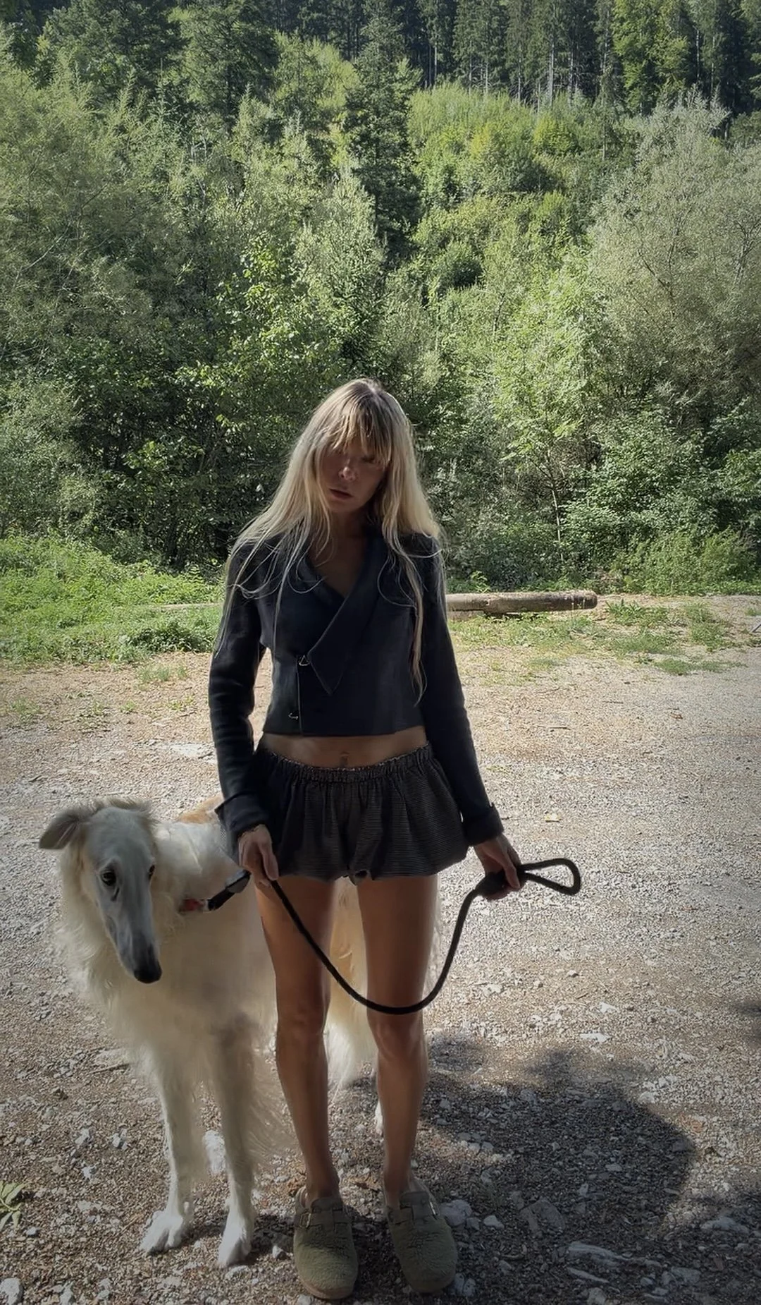 A young fashion woman with long blonde hair, wearing a grey cropped jacket and shorts, standing outdoors holding a leash connected to a white dog with a beige face and long fur.