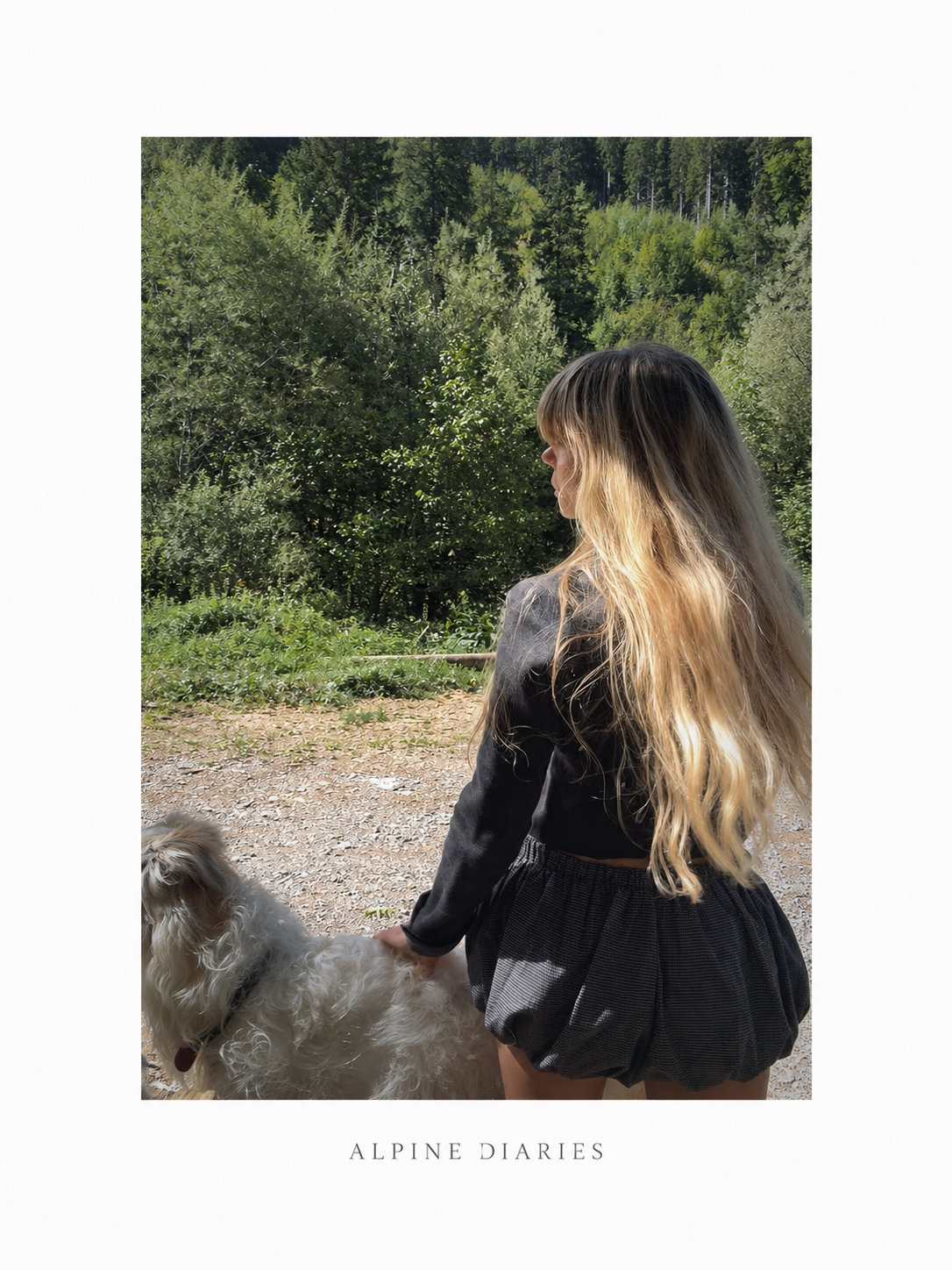 A young woman with long, wavy blonde hair wearing a woolen grey jacket and stylish fashion grey shorts, standing outdoors on a trail with green trees in the background, holding a big white dog by a leash.