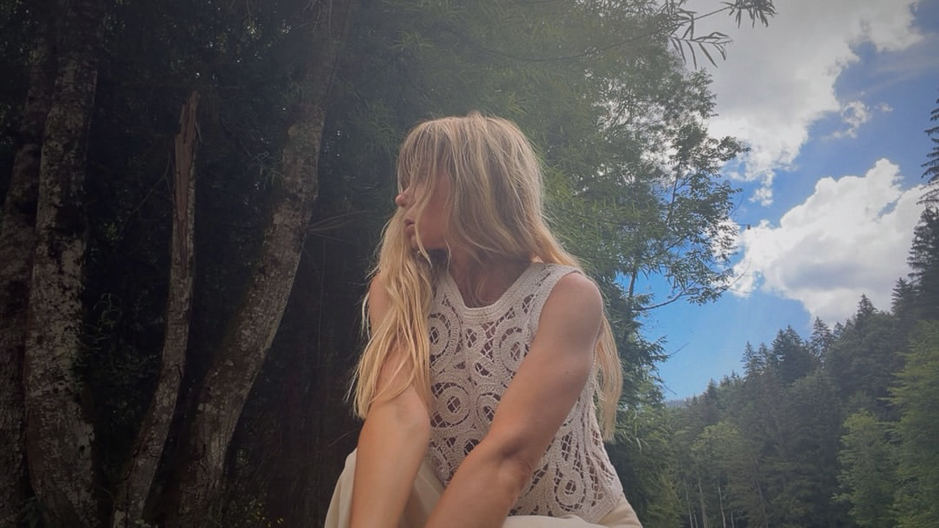 A woman with long blonde hair wearing a sleeveless lace top sitting outdoors near trees and a blue sky with clouds.
