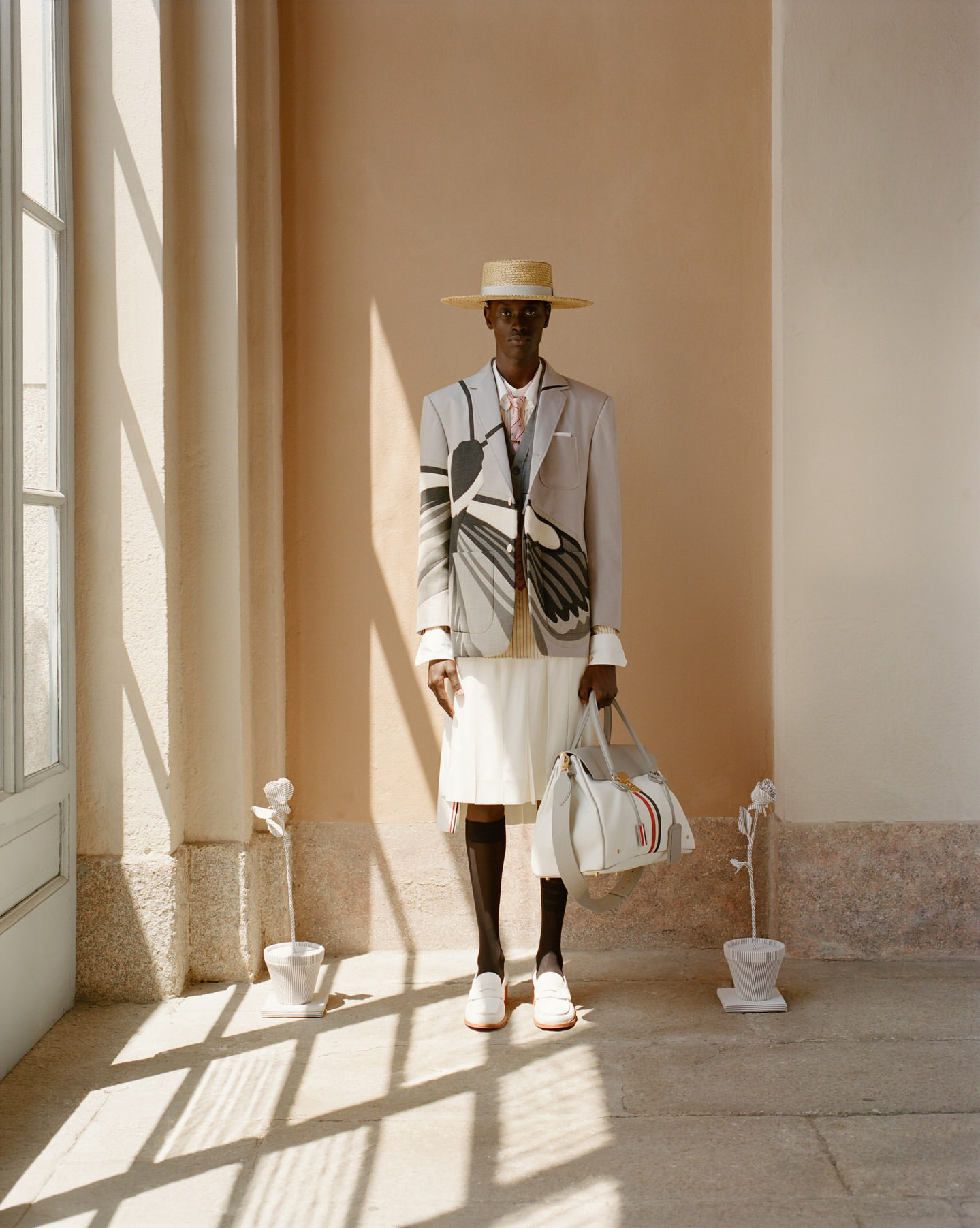 A person standing indoors near a window, dressed in a beige blazer with black and white abstract patterns, a white pleated skirt, and white sneakers. They are wearing a wide-brim straw hat, black stockings, and holding a large white handbag. The room has light-colored walls with sunlight casting shadows, and there are two small potted plants on the floor.