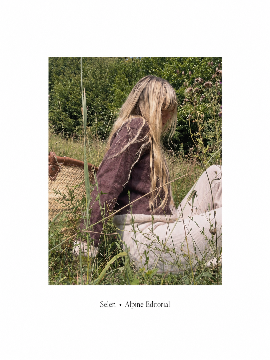 A young woman with long blonde hair sitting in tall grass outdoors, wearing a woolen Italian brown jacket and woolen light-colored milky pants, with a woven bag nearby, surrounded by greenery and wildflowers.