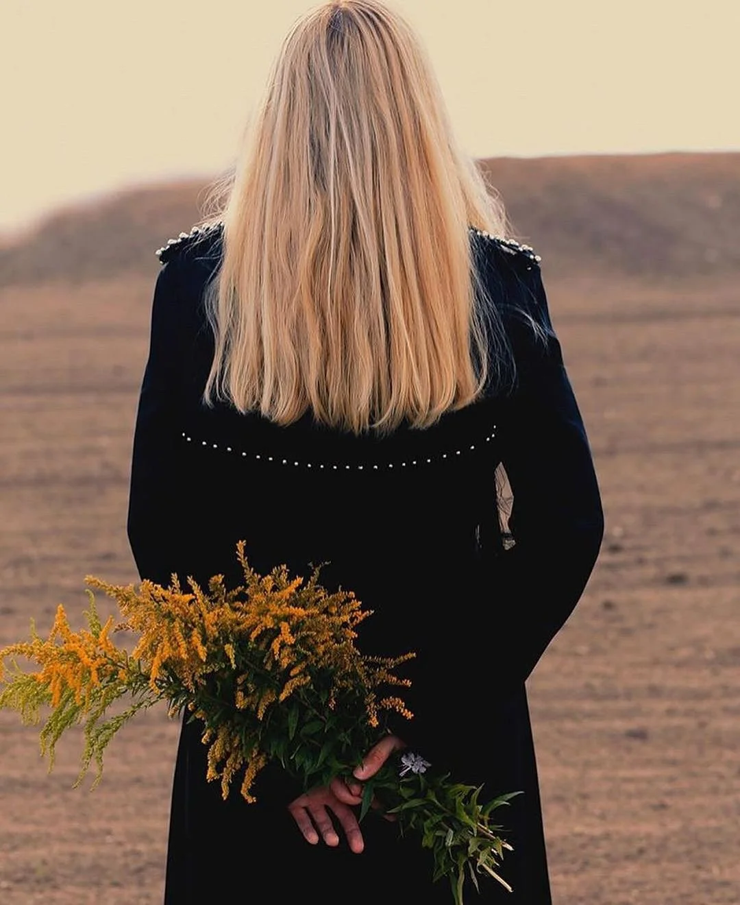 A woman with long blonde hair is standing with her back to the camera in a field, holding a bouquet of yellow flowers behind her back.