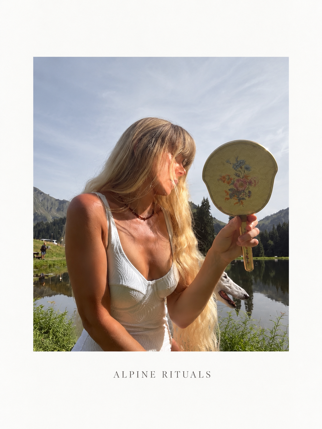A woman with long blonde hair wearing a white swimming suit looks at a sophisticated vintage floral hand mirror outdoors near a lake with mountains in the background.