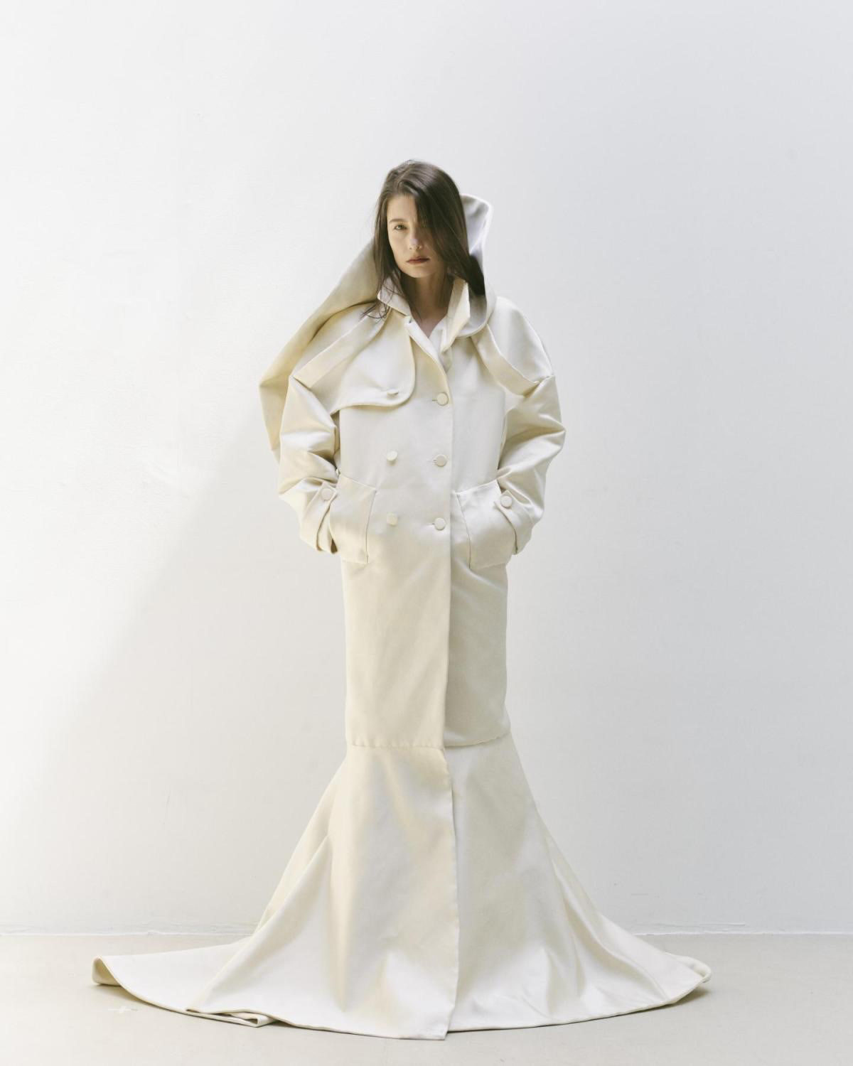 A woman wearing a long, cream-colored coat with a flared bottom, standing against a plain white background.