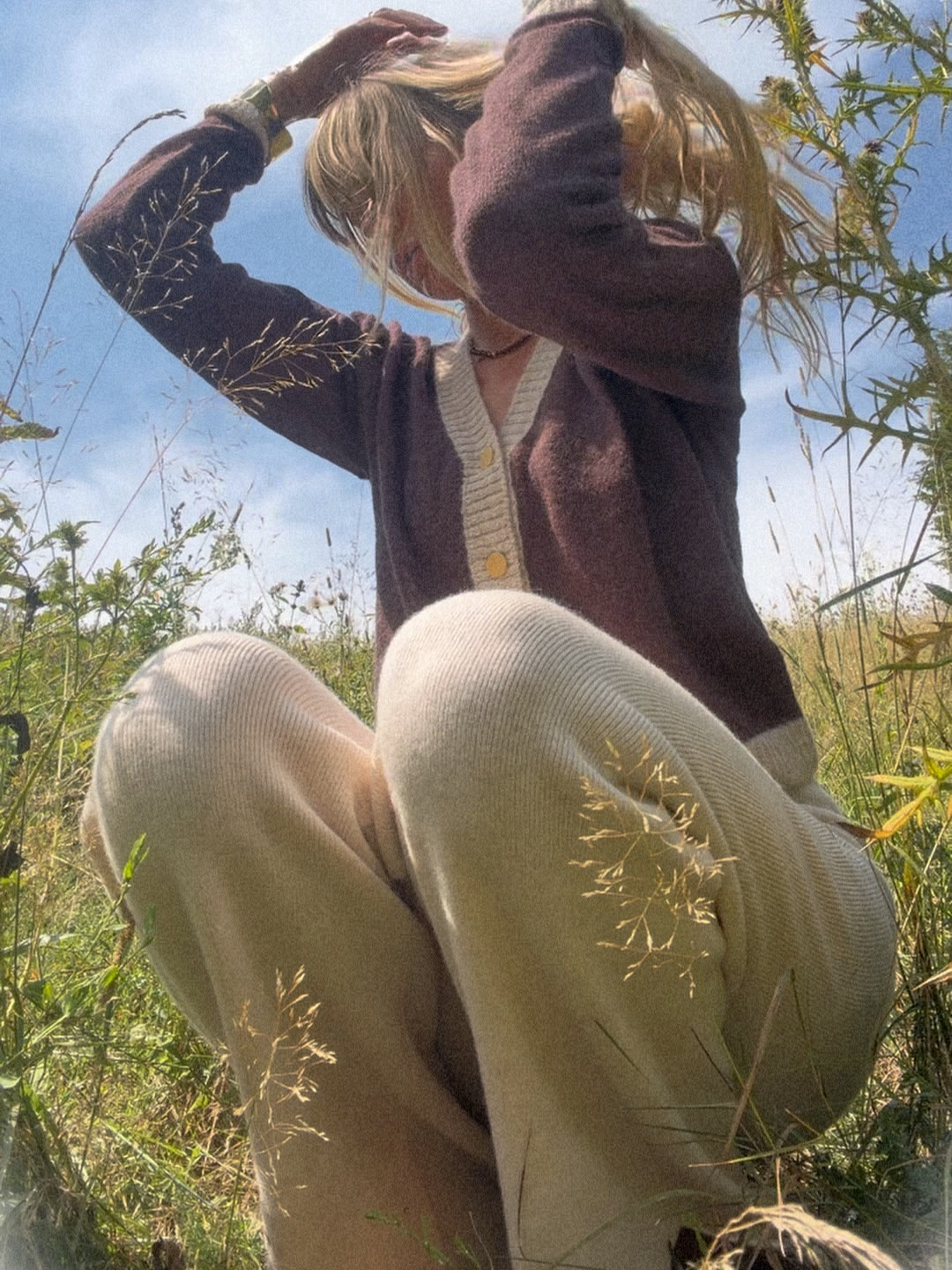 Charming girl sitting in a grassy field during summer daytime, wearing woolen beige pants and a brown cardigan with golden buttons, with hand in hair, under a partly cloudy sky.