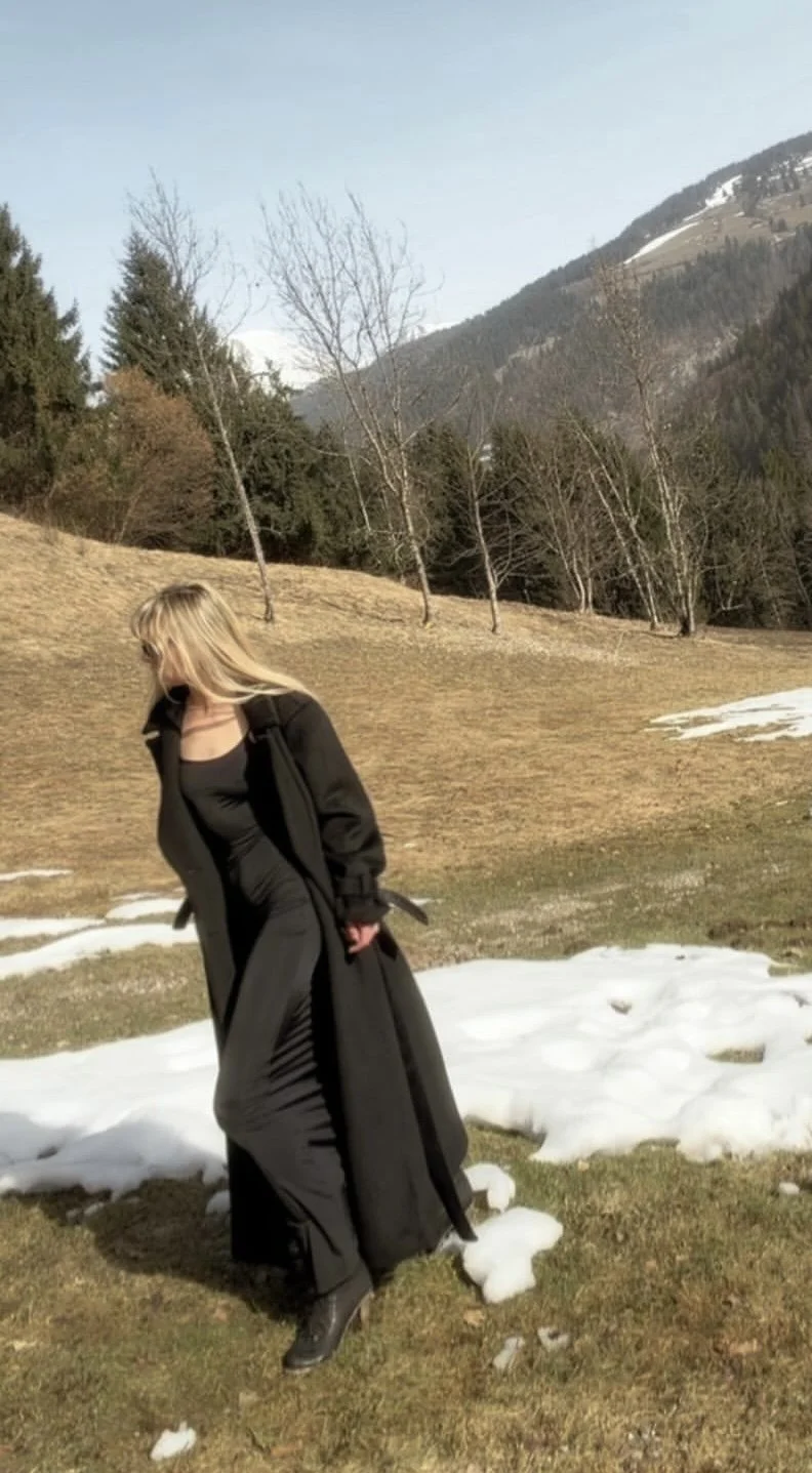A woman in a long black coat and dress standing outdoors on a grassy hillside with patches of snow, mountains and trees in the background on a clear day.