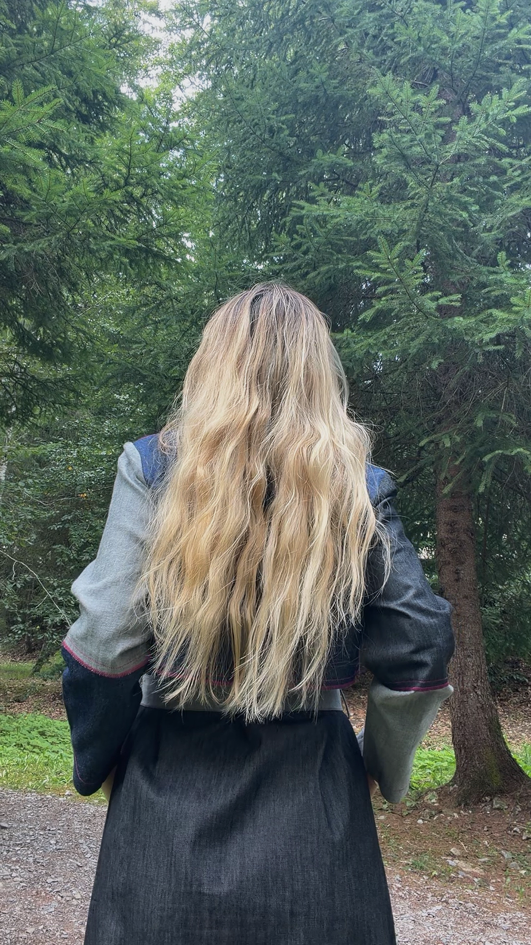 Back view of a girl with long, wavy blonde hair, wearing a jeans blue trench with grey and dark patchwork details, standing outdoors amidst green trees.