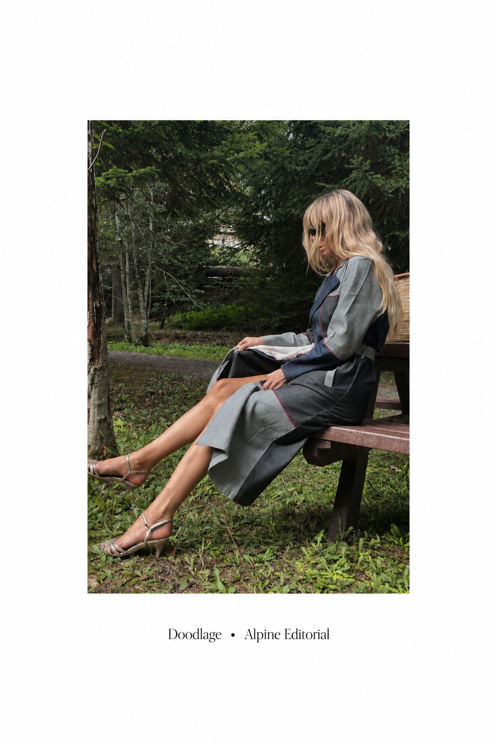 A woman with long blonde hair sitting on a wooden park bench in a wooded area, wearing a gray and black jeans patchwork trench with red piping and silver high heels.