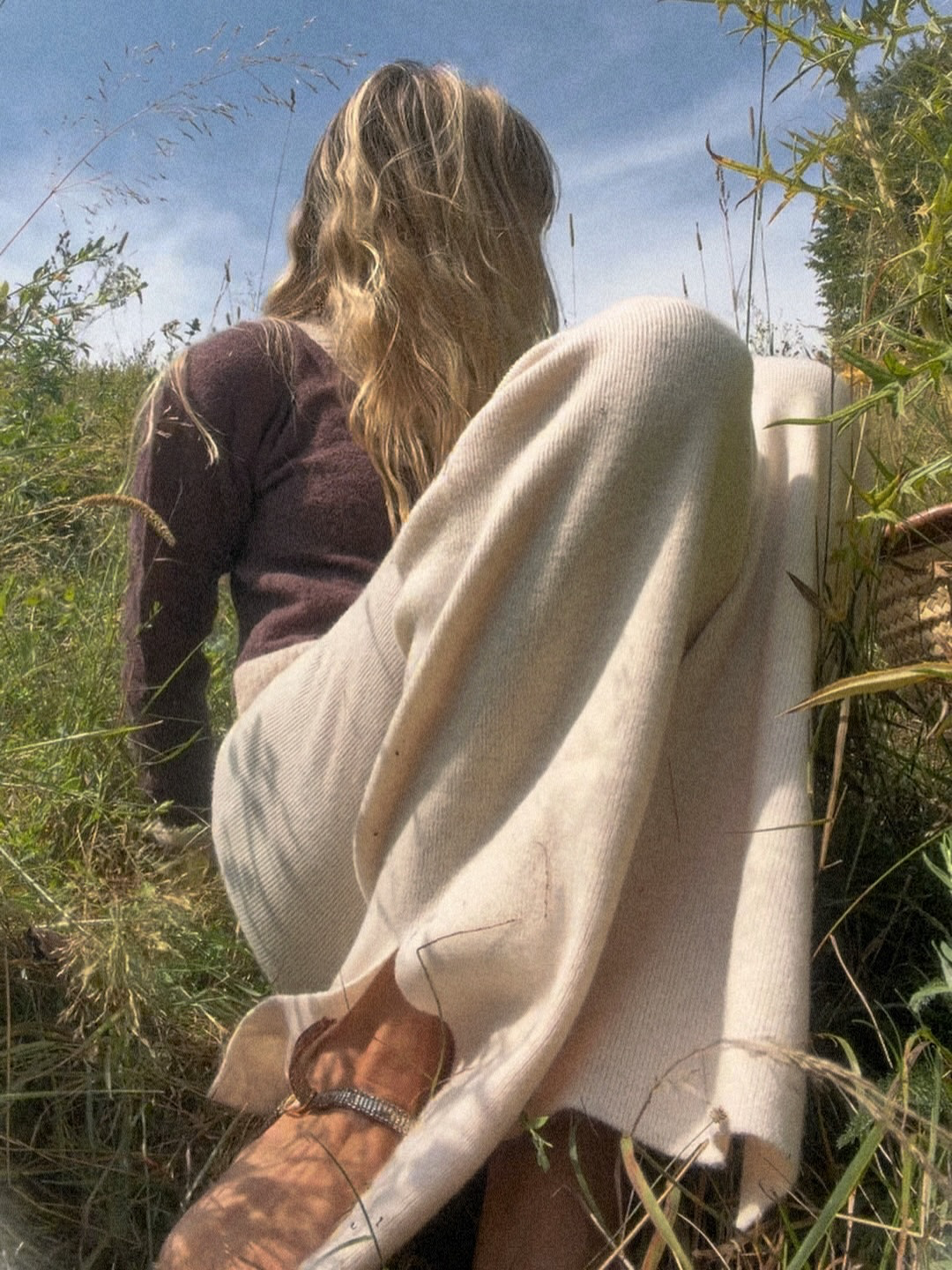 Model with long hair sitting outdoors in tall grass during daytime, wearing a white sweatshirt and white woolen pants, with a blue sky overhead.