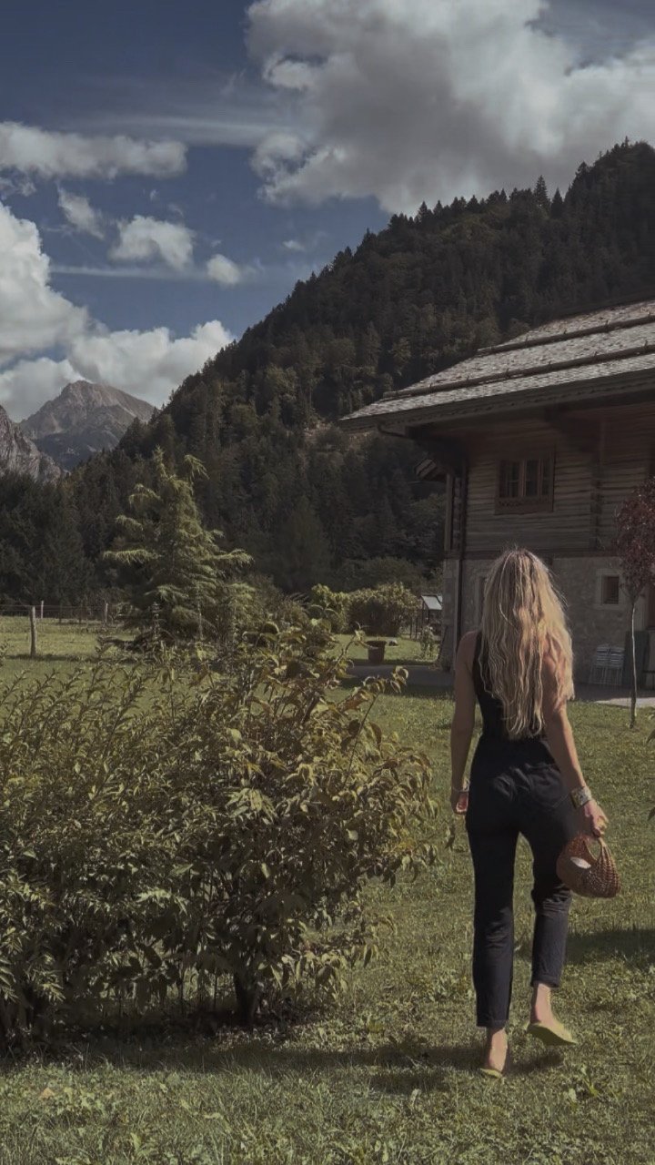 A woman with long blonde hair walking away on a grassy lawn holding a straw small bag, with an alpine chalet and a mountain landscape in the background, partly cloudy sky.