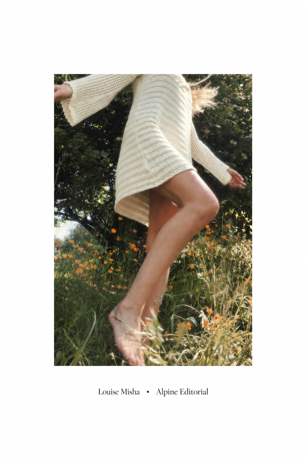 A model in a cream knitted dress standing in a field of orange flowers with greenery and trees in the background.