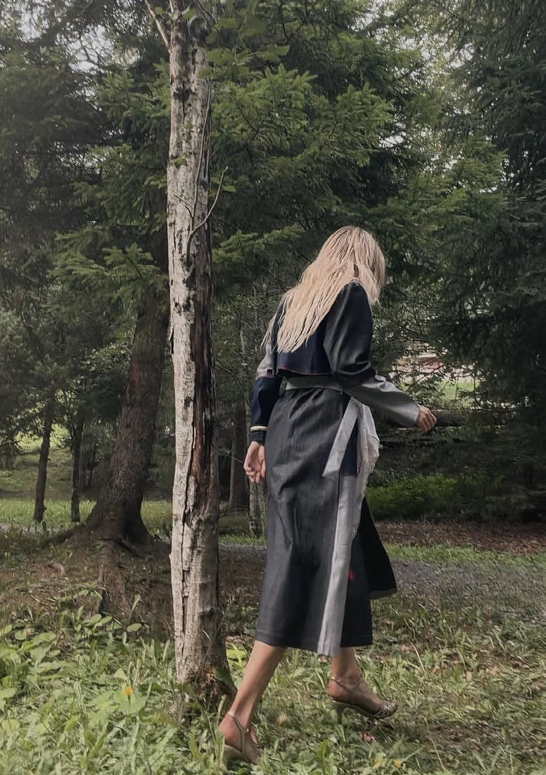 Woman with long blonde hair walking in a forest surrounded by trees and greenery, wearing a black and gray coat and high heels.