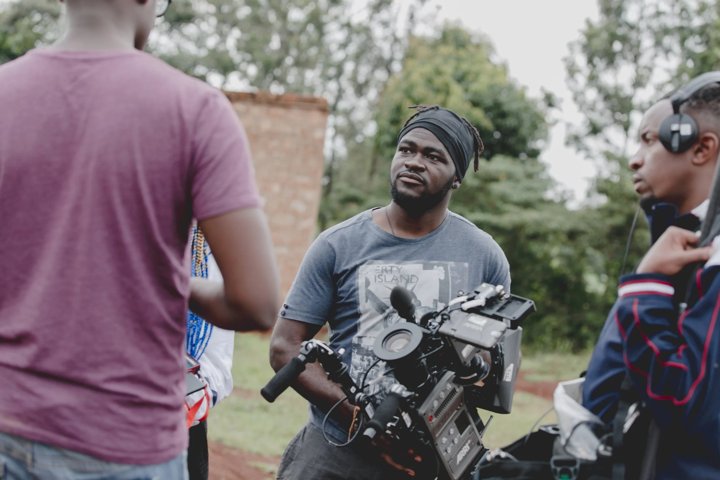 A man with a dark headband holding a professional video camera, surrounded by three other people outdoors with trees in the background.