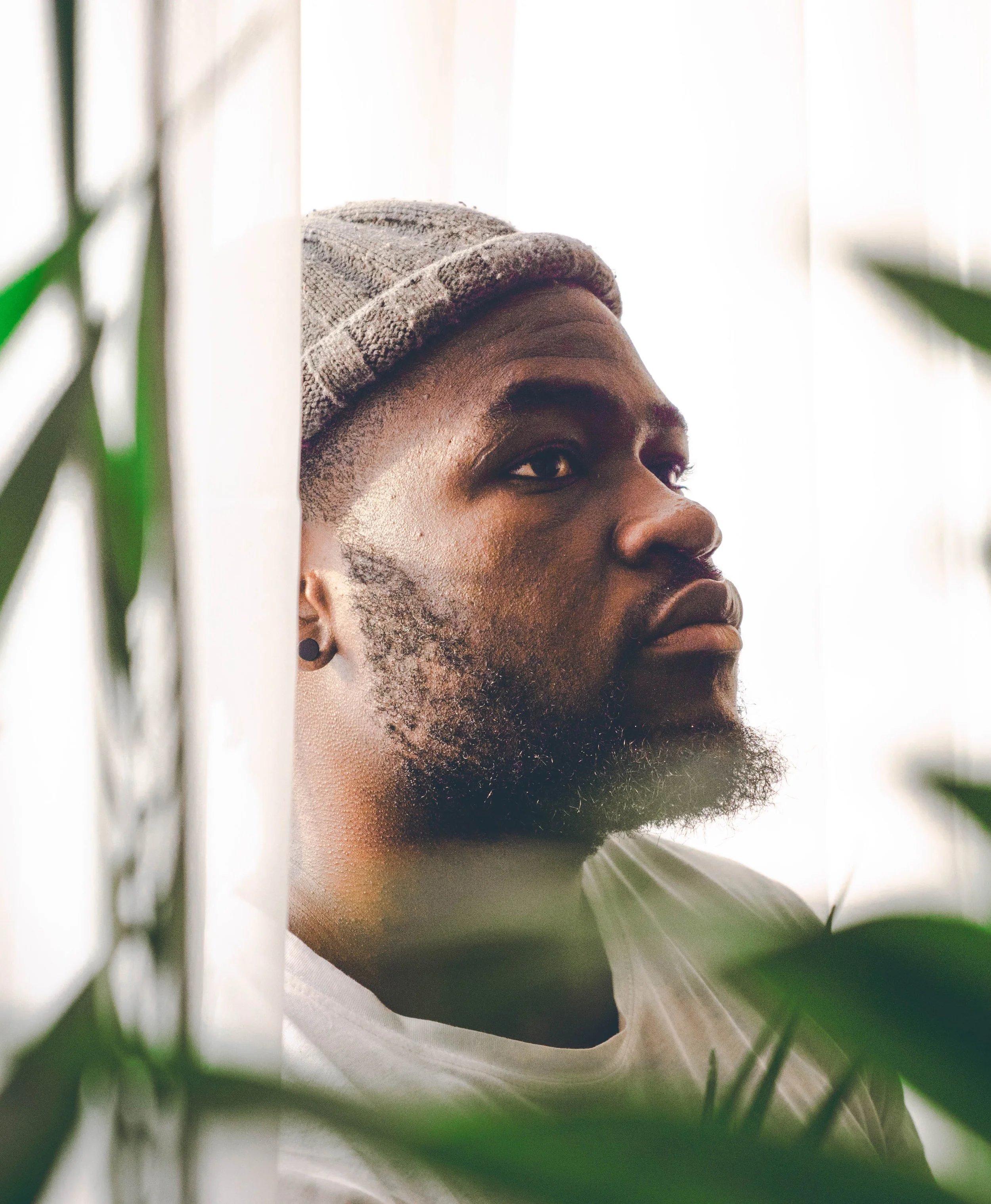 Close-up of a person's side profile with a beard, wearing a knit beanie, surrounded by green foliage, against a white background.