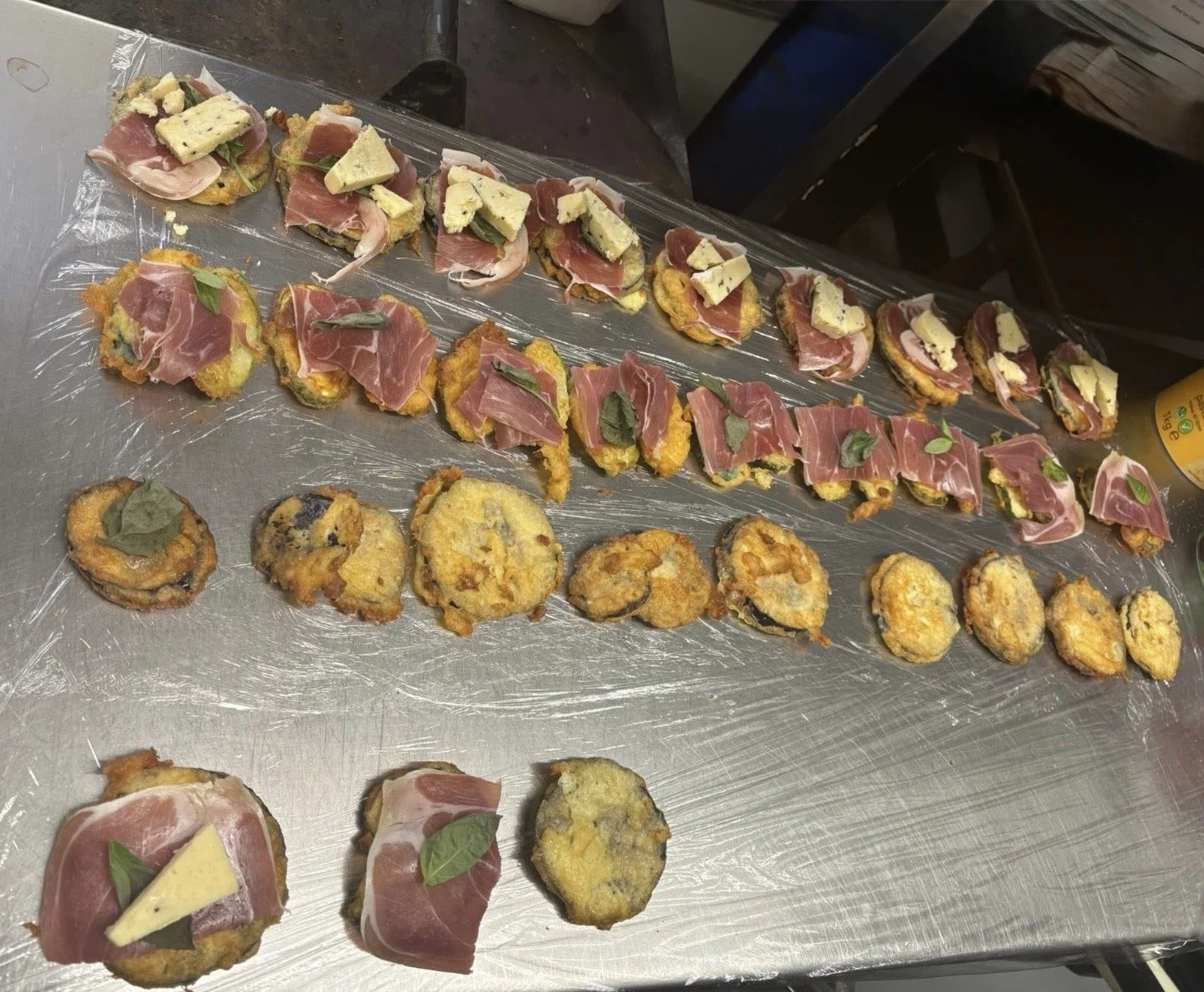 Assorted small appetizers on a metal tray, including slices of cured meat, cheese, and fried items with garnishes.